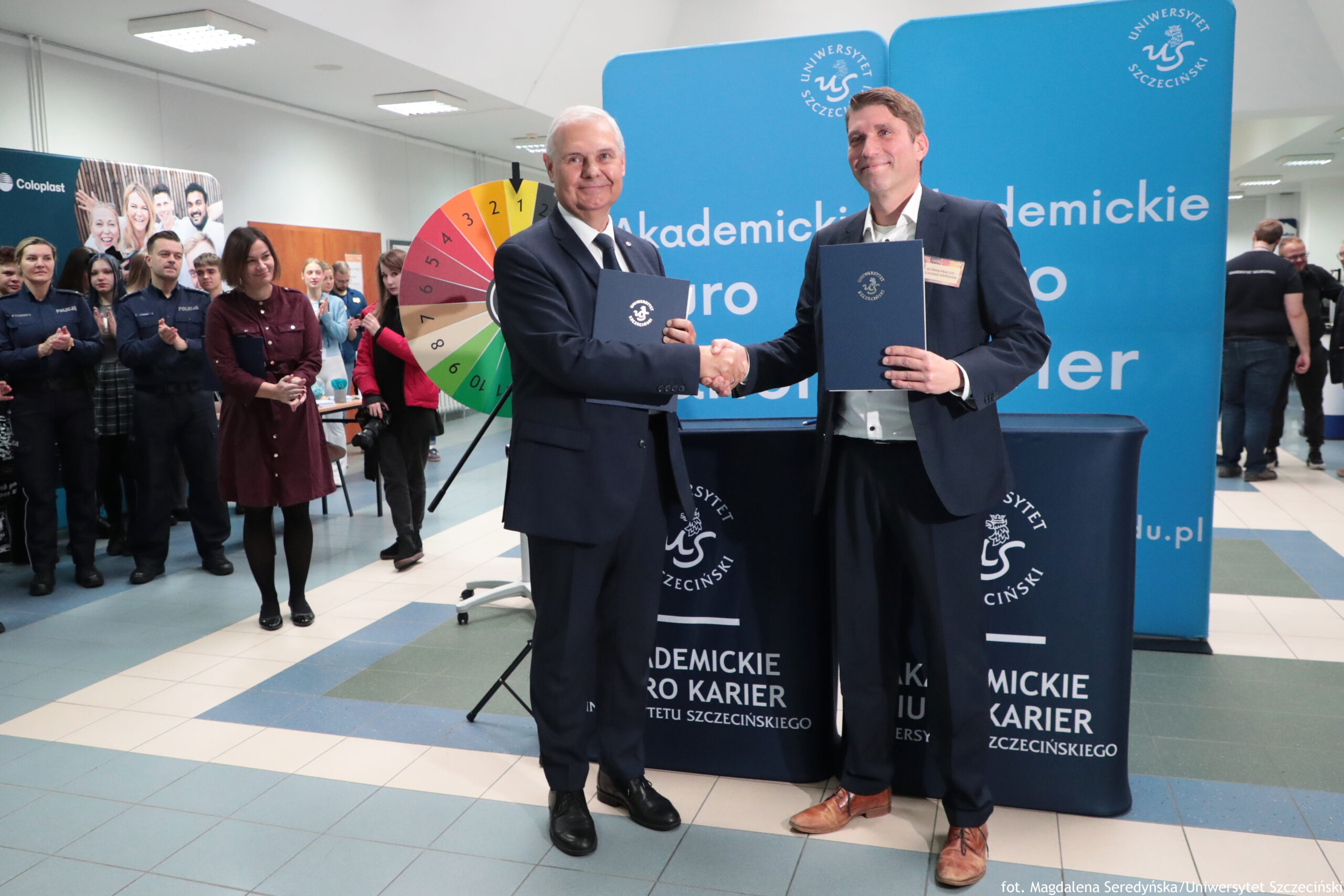 Dwóch mężczyzn w garniturach stojących na środku i podających sobie ręce, każdy trzyma granatową teczkę z godłem. Obaj są uśmiechnięci; mężczyzna po lewej ma siwe włosy, a mężczyzna po prawej brązowe włosy. W tle stoi duża niebieska ścianka z napisem „Uniwersytet Szczeciński” i dużym tekstem zawierającym słowa „Akademickie … Karier”. Przed ścianką jest stół przykryty granatowym obrusem z białym napisem „Akademickie Biuro Karier Uniwersytetu Szczecińskiego”. Po lewej stronie widać grupę osób obserwujących i klaszczących. Obok nich stoi kolorowe koło losujące z polami w różnych kolorach i numerami.
