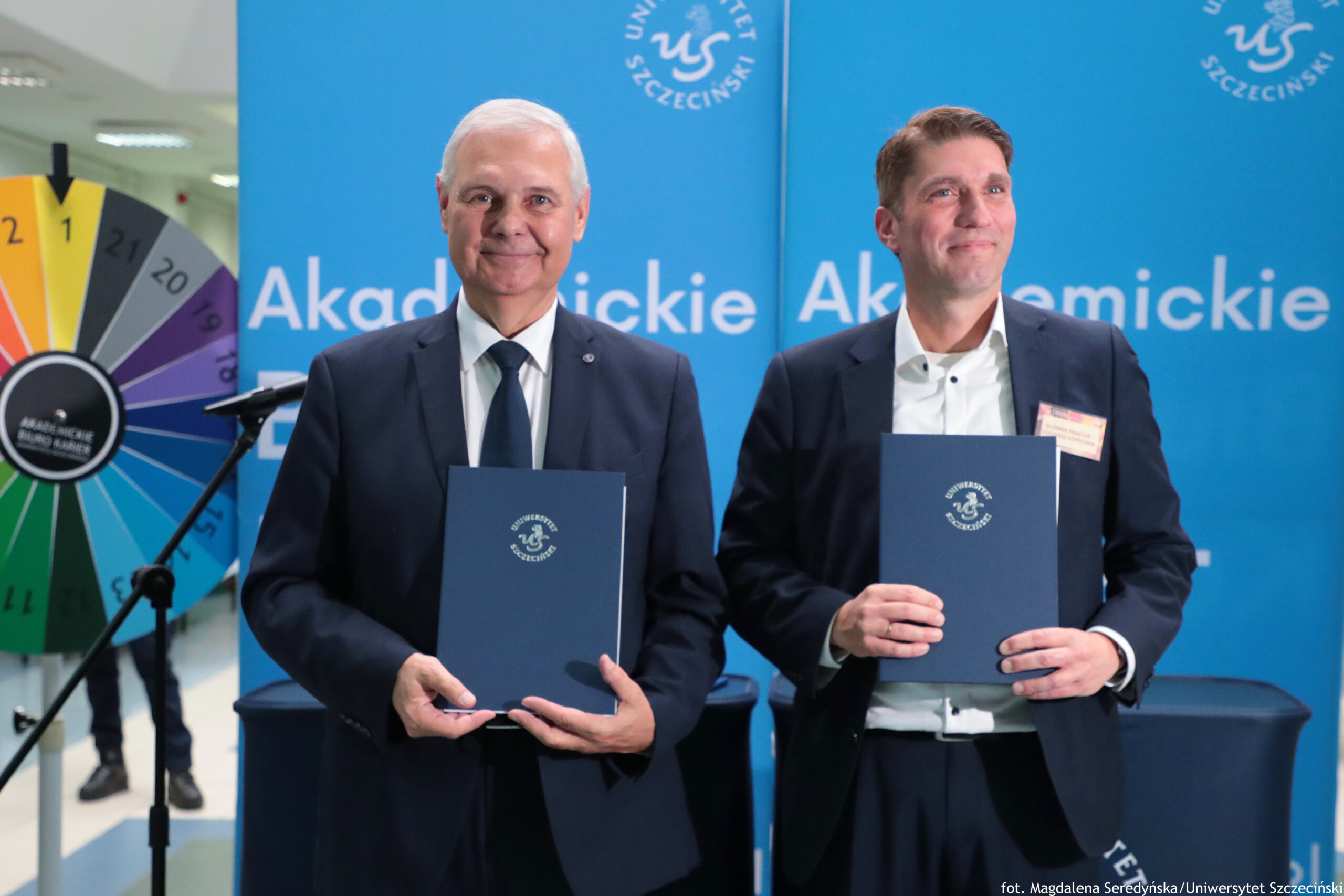 Dwóch mężczyzn w garniturach stojących przed niebieskim tłem z napisem „Akademickie” i logotypem „Uniwersytet Szczeciński”, trzymających granatowe teczki z godłem uczelni. Mężczyzna po lewej ma siwe włosy, białą koszulę i ciemny krawat; uśmiecha się do aparatu. Mężczyzna po prawej ma krótkie brązowe włosy, białą koszulę bez krawata i identyfikator przypięty do marynarki; patrzy lekko w bok i też się uśmiecha. Po lewej stronie kadru widać kolorowe koło z numerami (jak ruletka/koło fortuny) oraz stojak z mikrofonem. Na dole po prawej jest podpis: „fot. Magdalena Sieredyńska/Uniwersytet Szczeciński”.