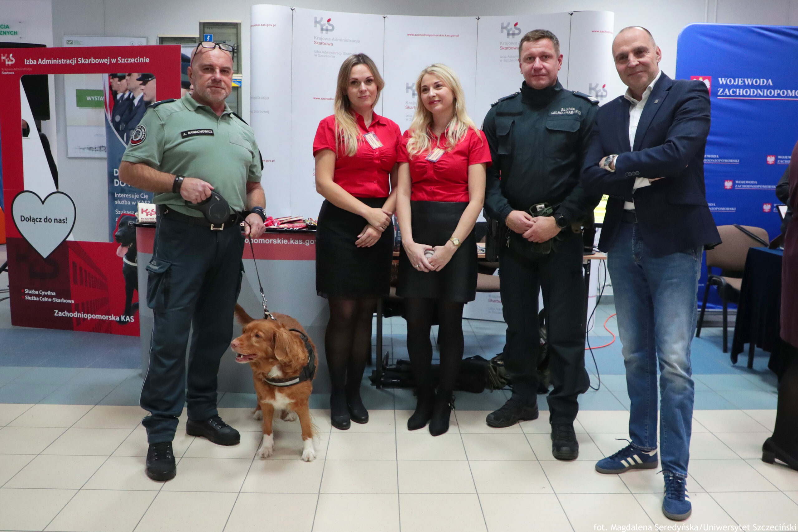 Grupa pięciu dorosłych osób stojących w pomieszczeniu na tle stoisk informacyjnych, z psem na smyczy na pierwszym planie. Od lewej stoi mężczyzna w zielonym mundurze, obok niego na podłodze średniej wielkości brązowo-biały pies w uprzęży. W środku stoją dwie kobiety w czerwonych koszulach i czarnych spódnicach. Po prawej stoi mężczyzna w czarnym mundurze. Skrajnie po prawej stoi mężczyzna w granatowej marynarce i jasnej koszuli, z założonymi rękami. W tle widać banery i stoiska z logotypami oraz czytelnymi fragmentami tekstu: „Izba Administracji Skarbowej w Szczecinie”, „Dołącz do nas!”, „Zachodniopomorska KAS”, a na niebieskim banerze „WOJEWODA ZACHODNIOPOMORSKI”. Na dole zdjęcia jest podpis: „fot. Magdalena Seredyńska / Uniwersytet Szczeciński”.