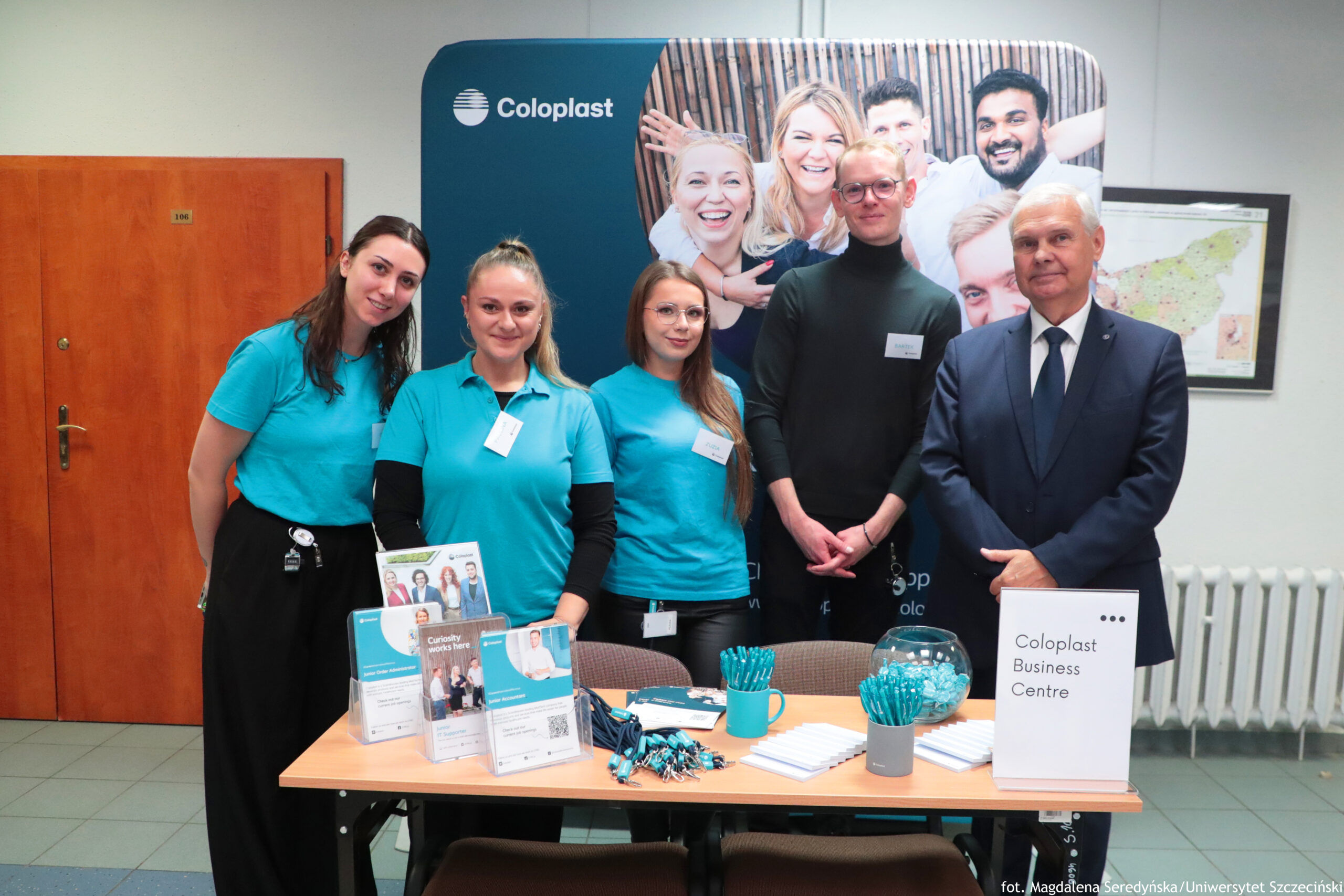 Grupa pięciu osób stojących za stołem promocyjnym firmy Coloplast. Na stole leżą stojaki z ulotkami, smycze z identyfikatorami, długopisy w pojemnikach, stos białych notesów lub karteczek oraz szklana misa z turkusowymi drobiazgami. Po prawej stronie na stole stoi tabliczka z napisem „Coloplast Business Centre”. Trzy kobiety mają na sobie turkusowe koszulki polo i identyfikatory; stoją po lewej stronie. Obok nich stoi mężczyzna w ciemnym golfie i okularach z identyfikatorem. Po prawej stoi starszy mężczyzna w granatowym garniturze, białej koszuli i krawacie. W tle znajduje się duża ścianka reklamowa z logo „Coloplast” i zdjęciem uśmiechniętych osób. Po lewej widać drzwi, a po prawej fragment mapy na ścianie i grzejnik. Na dole zdjęcia jest mały podpis: „fot. Magdalena Seredyńska / Uniwersytet Szczeciński”.