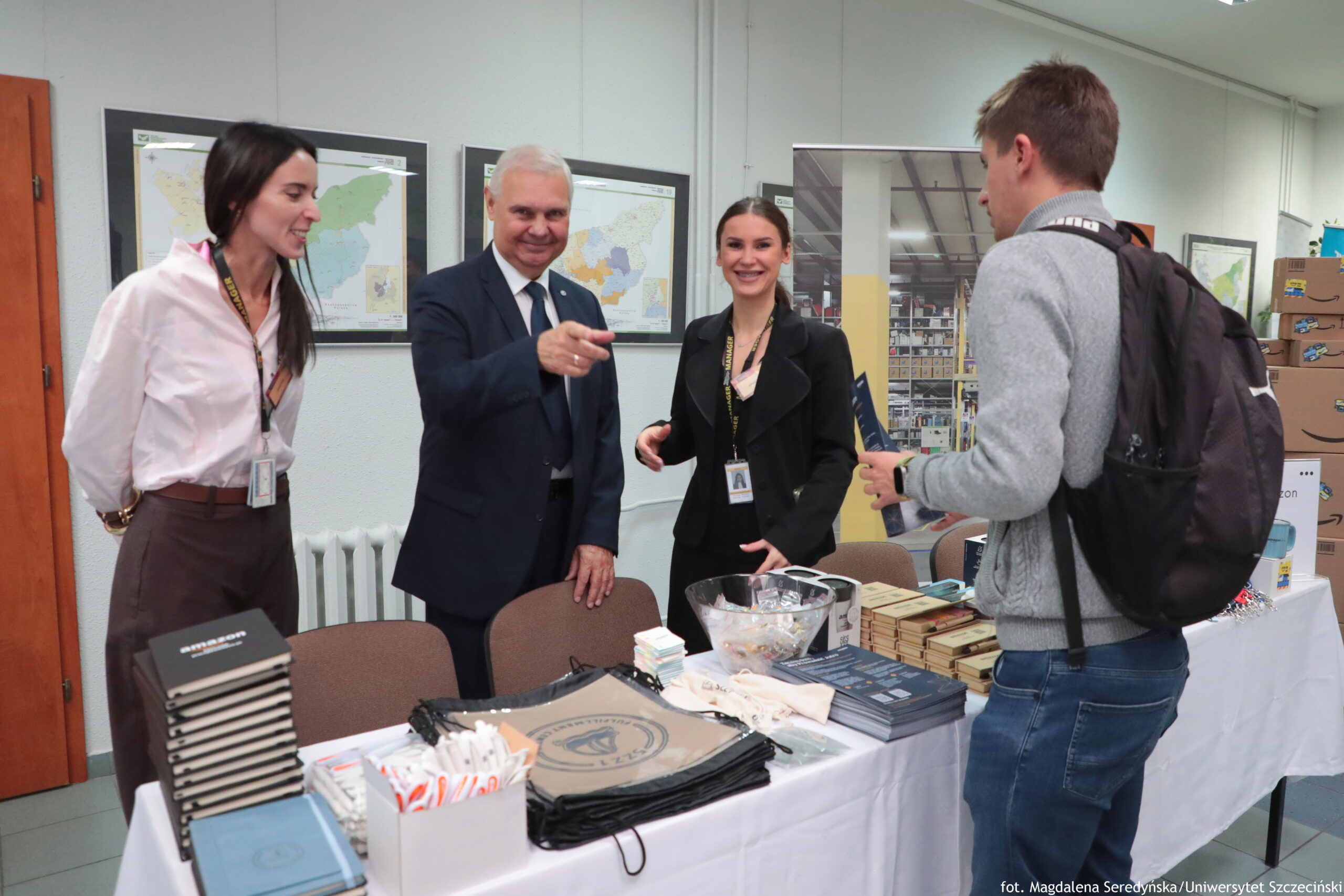 Stół informacyjny z materiałami promocyjnymi i czterema osobami stojącymi dookoła w pomieszczeniu biurowym. Na pierwszym planie stoi długi stół przykryty białym obrusem. Na blacie leżą stosy książek lub notesów, kilka stert broszur, pudełko z długopisami, przezroczysta misa z drobnymi upominkami oraz czarne worki materiałowe z nadrukiem. Za stołem stoją trzy osoby w identyfikatorach na smyczach: po lewej kobieta z długimi ciemnymi włosami w białej koszuli i brązowych spodniach; pośrodku starszy mężczyzna o siwych włosach w granatowym garniturze i krawacie, z uśmiechem, wyciągający rękę i wskazujący palcem w stronę osoby po prawej; obok niego młoda kobieta z ciemnymi włosami upiętymi do tyłu, w czarnym stroju, uśmiechnięta. Po prawej stronie, przodem do nich, stoi młody mężczyzna w szarym swetrze i dżinsach, z czarnym plecakiem; trzyma w ręku jakiś materiał (prawdopodobnie broszurę lub folder).