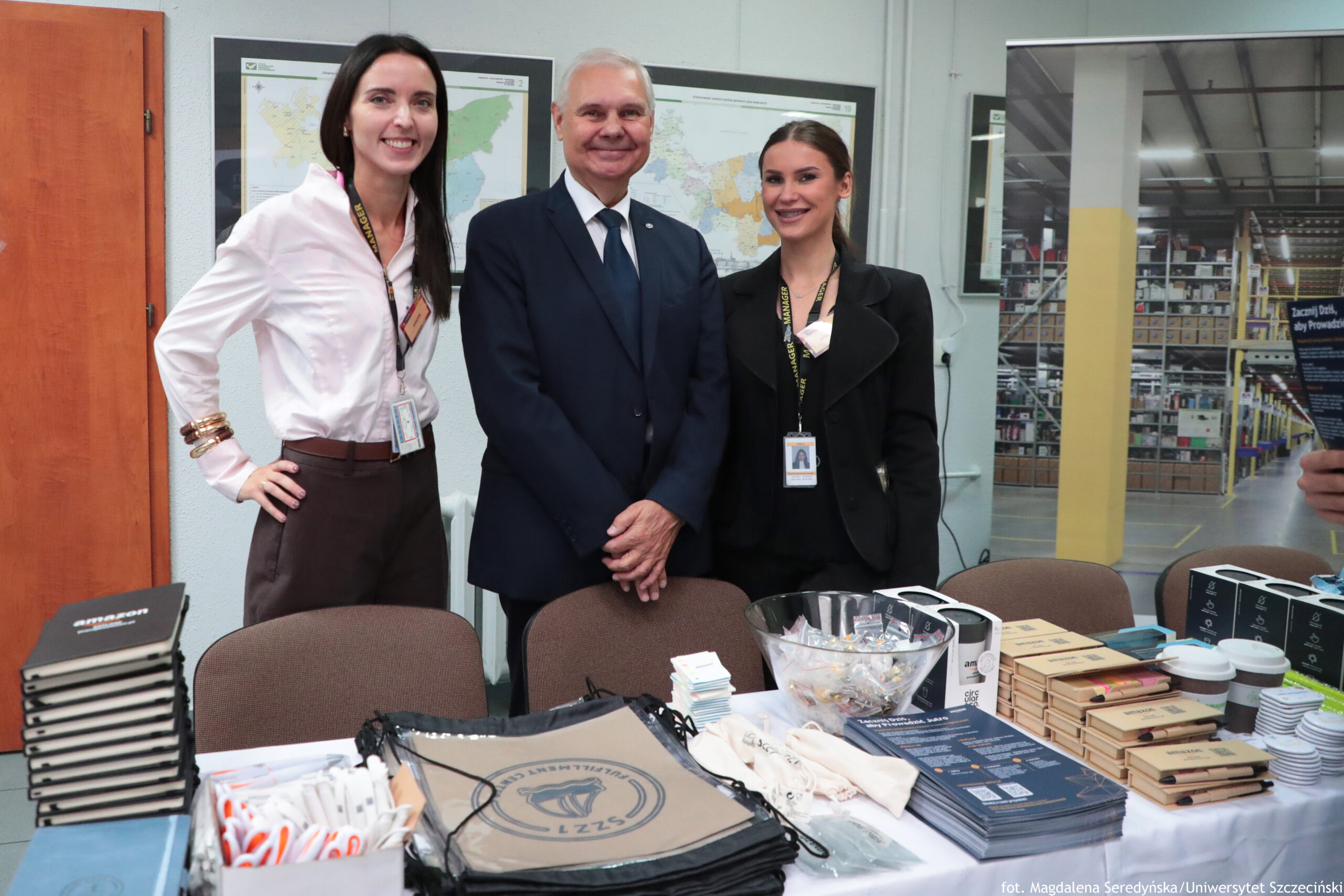 Trzy osoby pozujące do zdjęcia, stojące za stołem z materiałami promocyjnymi. Po lewej stoi kobieta o jasnej karnacji, długich ciemnych włosach, w białej koszuli i brązowych spodniach; ma identyfikator na smyczy i bransoletki na nadgarstku, uśmiecha się. W środku stoi starszy mężczyzna o jasnej karnacji i siwych włosach, w ciemnym garniturze z krawatem; trzyma dłonie złożone z przodu i uśmiecha się. Po prawej stoi kobieta o jasnej karnacji, z ciemnymi włosami związanymi z tyłu, w czarnym stroju; ma identyfikator na smyczy i uśmiecha się. Na pierwszym planie jest stół przykryty białym obrusem z wieloma rzeczami: stosy notesów lub zeszytów, długopisy, smyczki, ulotki, kubki papierowe, pudełka i miska z drobnymi przedmiotami. W tle wiszą mapy na ścianie, a po prawej stronie widać otwartą przestrzeń magazynu z regałami. W prawym dolnym rogu jest podpis fotograficzny: „fot. Magdalena Seredyńska / Uniwersytet Szczeciński”.