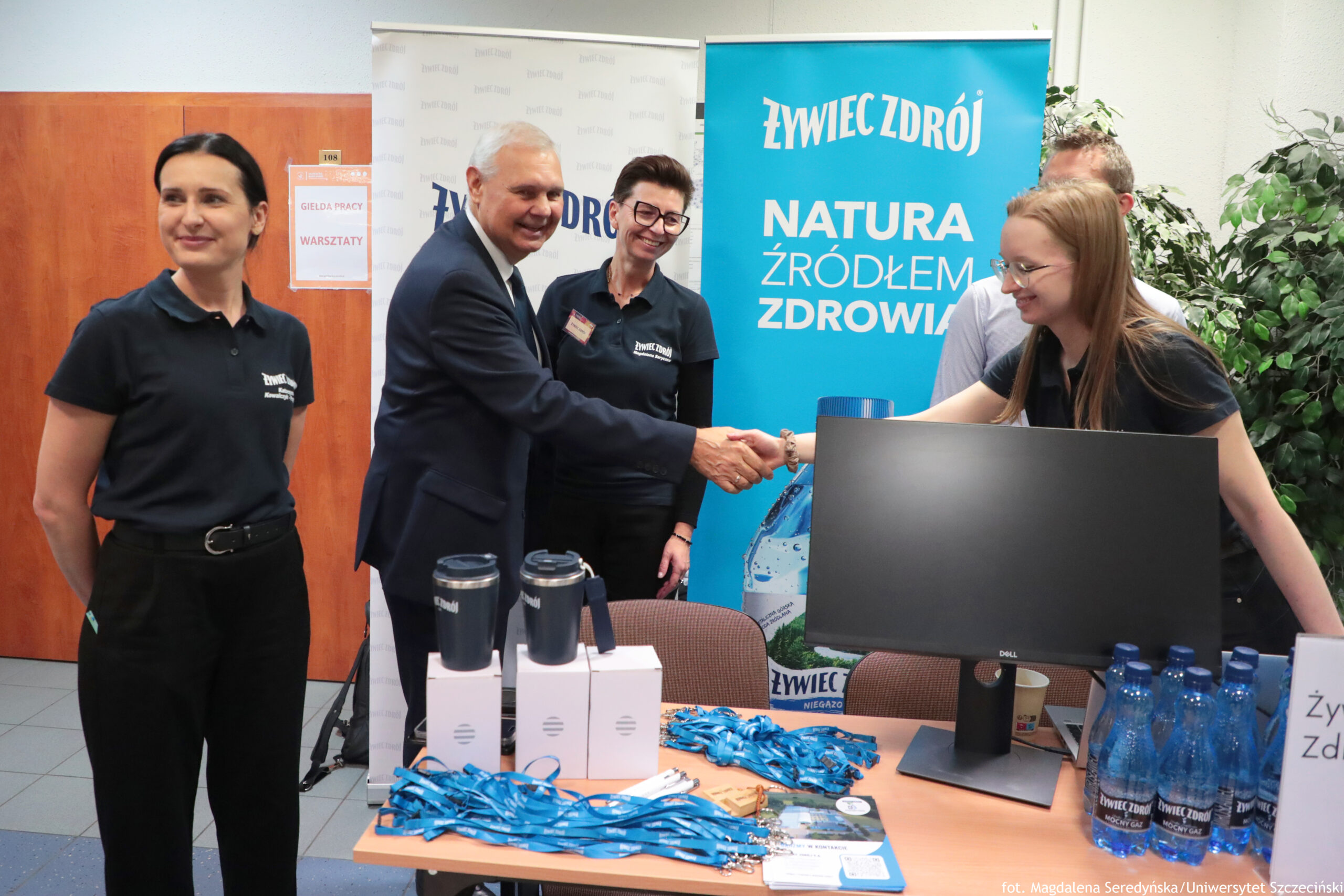 Stoisko promocyjne marki Żywiec Zdrój na wydarzeniu, z kilkoma osobami stojącymi wokół stołu. W centrum starszy mężczyzna o siwych włosach w ciemnym garniturze ściska dłoń młodej kobiety z długimi jasnorudymi włosami i okularami; kobieta pochyla się lekko nad stołem i uśmiecha się. Obok nich stoi kobieta w czarnej koszulce polo z logo Żywiec Zdrój, z krótkimi ciemnymi włosami i okularami, także uśmiechnięta. Po lewej stronie kadru stoi kolejna kobieta w czarnej koszulce polo z logo, z ciemnymi włosami związanymi z tyłu, patrząca w bok. W tle z prawej widać jeszcze fragment postaci mężczyzny w białej koszuli. Za stoiskiem znajduje się duży niebieski baner z napisem: „ŻYWIEC ZDRÓJ” oraz „NATURA ŹRÓDŁEM ZDROWIA”. Na stole leżą niebieskie smycze, stoją dwa ciemne kubki termiczne na białych pudełkach, widoczny jest monitor komputerowy marki Dell, a po prawej stronie kilka niebieskich butelek wody Żywiec Zdrój. W tle po lewej na drzwiach wisi kartka z napisem „GIEŁDA PRACY” oraz „WARSZTATY”.