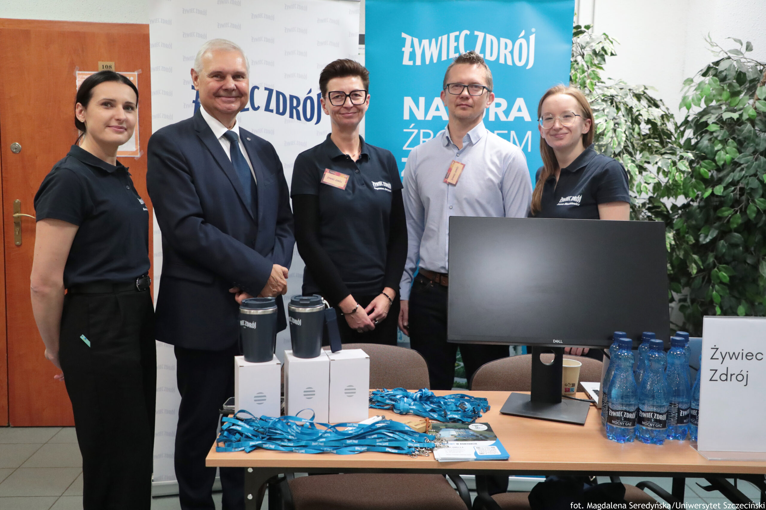 Grupa pięciu osób stojących za stołem promocyjnym w pomieszczeniu. Trzy kobiety i dwóch mężczyzn pozują do zdjęcia; cztery osoby mają na sobie czarne koszulki polo z logo, jedna osoba (mężczyzna) ma jasną koszulę, a drugi mężczyzna ma ciemny garnitur z krawatem. W tle znajdują się roll-upy z napisem „ŻYWIEC ZDRÓJ” oraz roślina doniczkowa. Na stole leżą niebieskie smycze z identyfikatorami, kilka białych pudełek, dwa czarne kubki/termosy, monitor komputerowy marki Dell, oraz zgrzewka małych butelek wody z etykietą „Żywiec Zdrój”. Z prawej strony stoi kartka/oznakowanie z napisem „Żywiec Zdrój”. W prawym dolnym rogu widoczny jest podpis: „fot. Magdalena Seredyńska / Uniwersytet Szczeciński”.