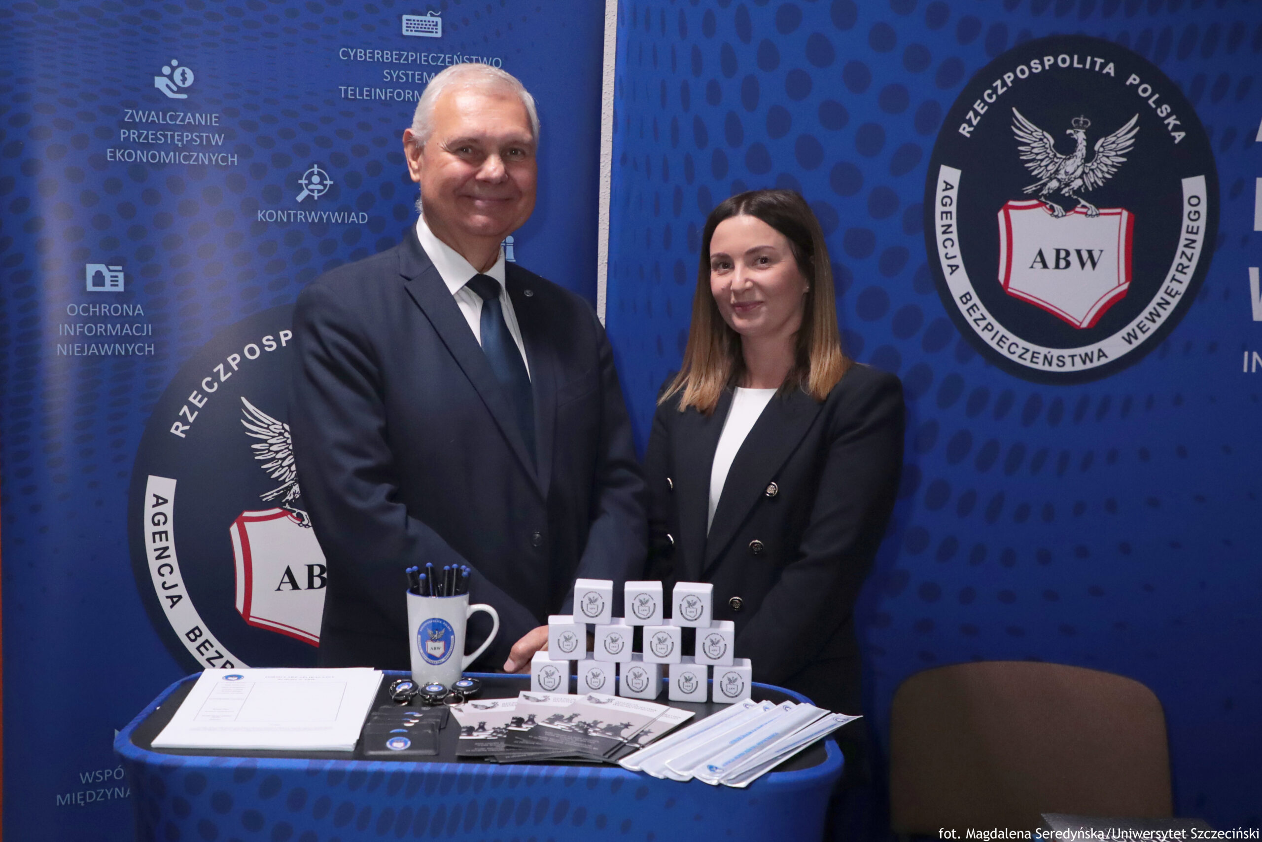 Dwie osoby stojące za stołem na tle niebieskich banerów z godłem i napisem „RZECZPOSPOLITA POLSKA” oraz „AGENCJA BEZPIECZEŃSTWA WEWNĘTRZNEGO” i skrótem „ABW”. Po lewej stronie stoi mężczyzna o jasnej skórze, siwych włosach, w ciemnym garniturze, białej koszuli i ciemnym krawacie; uśmiecha się. Po prawej stronie stoi kobieta o jasnej skórze, prostych włosach do ramion (ciemniejsze u góry, jaśniejsze na końcach), w ciemnej marynarce i jasnej bluzce; także lekko się uśmiecha. Na stole leżą materiały promocyjne: kubek z długopisami, kilka okrągłych przypinek lub breloków, ulotki, dokumenty oraz stos jednorazowych maseczek. Na środku stoi ułożona piramidka z małych białych pudełek z nadrukowanym okrągłym logo. W prawym dolnym rogu zdjęcia jest podpis: „fot. Magdalena Seredyńska/Uniwersytet Szczeciński”.
