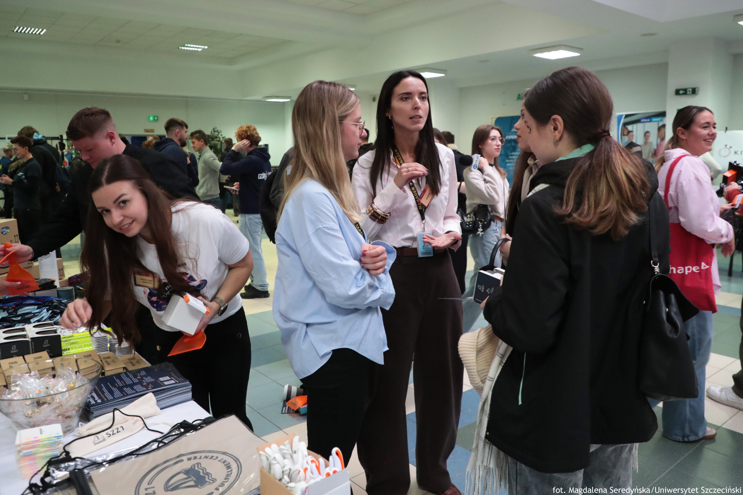 Grupa ludzi w holu, wydarzenia promocyjnego, przy stoisku z gadżetami. Widać kilka młodych dorosłych osób rozmawiających i stojących w tłumie. Na pierwszym planie po lewej stronie kobieta z długimi ciemnymi włosami w białej koszulce pochyla się nad stołem i trzyma małe białe pudełko; na stole leżą różne materiały i upominki, m.in. smycze, ulotki lub broszury, pudełka oraz pojemnik z drobnymi przedmiotami. W centrum stoją trzy kobiety w rozmowie: jedna w jasnoniebieskiej koszuli i okularach, obok niej kobieta w białej bluzce z identyfikatorem na smyczy, gestykulująca ręką, oraz kobieta zwrócona tyłem w ciemnej kurtce z torbą na ramieniu. W tle widać więcej uczestników, stoiska oraz plakaty. Na dole po prawej jest podpis: „fot. Magdalena Seredyńska/Uniwersytet Szczeciński”.