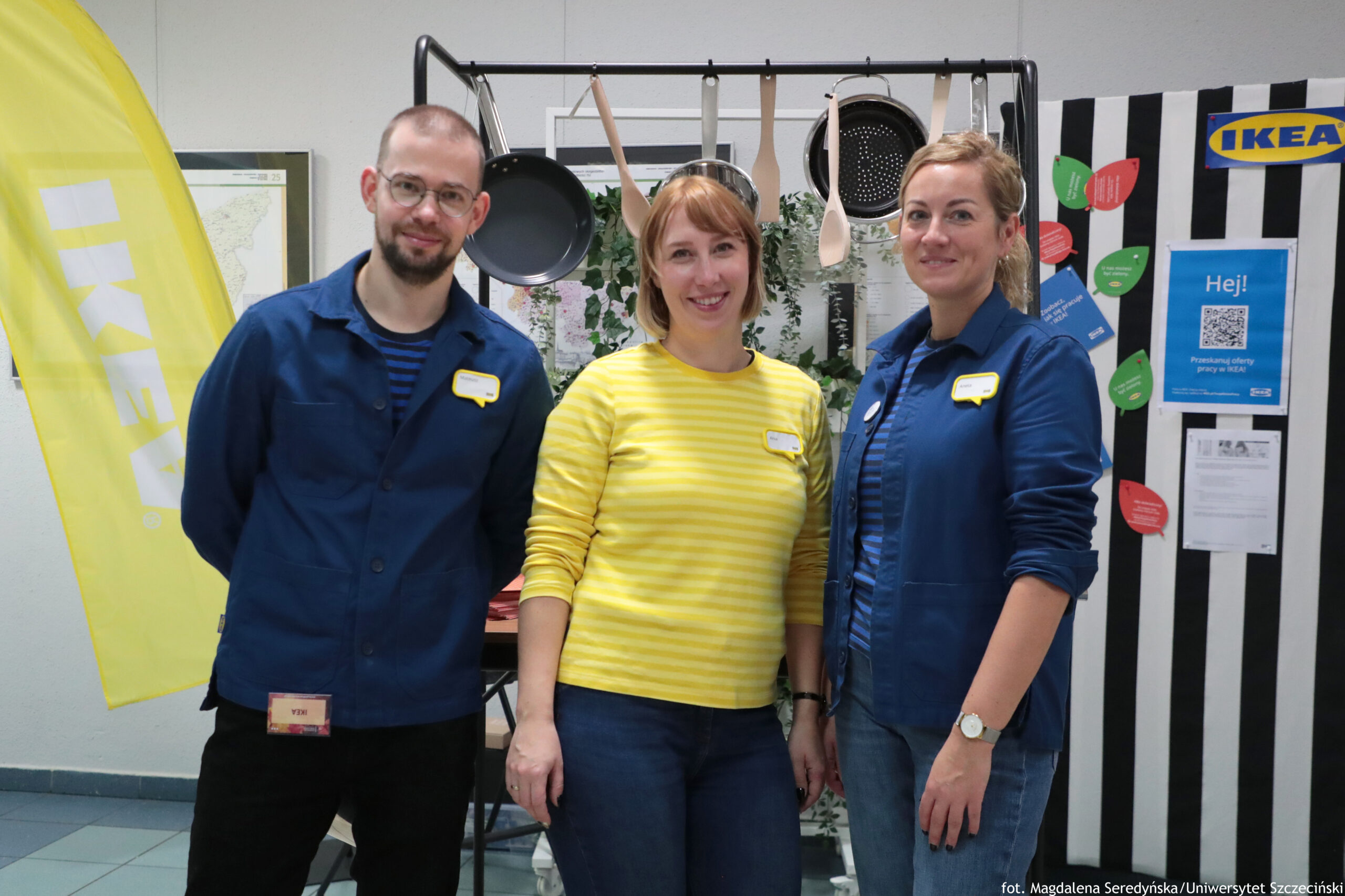 Trzy osoby stojące obok siebie w pomieszczeniu, pozujące do zdjęcia. Po lewej stoi mężczyzna o jasnej skórze, w okularach, z krótkimi włosami i krótką brodą; ma granatową kurtkę i identyfikator. W środku stoi kobieta o jasnej skórze, z jasnorudymi włosami do ramion; ma żółto-białą, pasiastą bluzkę i identyfikator. Po prawej stoi kobieta o jasnej skórze, z blond włosami upiętymi z tyłu; ma granatową kurtkę, identyfikator i zegarek na ręku. W tle wisi stelaż z kuchennymi przyborami (patelnie, łyżki, cedzak). Po lewej stronie kadru widać żółtą flagę z napisem IKEA. Po prawej jest tablica w czarno-białe pasy z logo IKEA oraz plakat z dużym napisem „Hej!” i kodem QR. Na dole po prawej stronie widać podpis: „fot. Magdalena Seredyńska/Uniwersytet Szczeciński”.