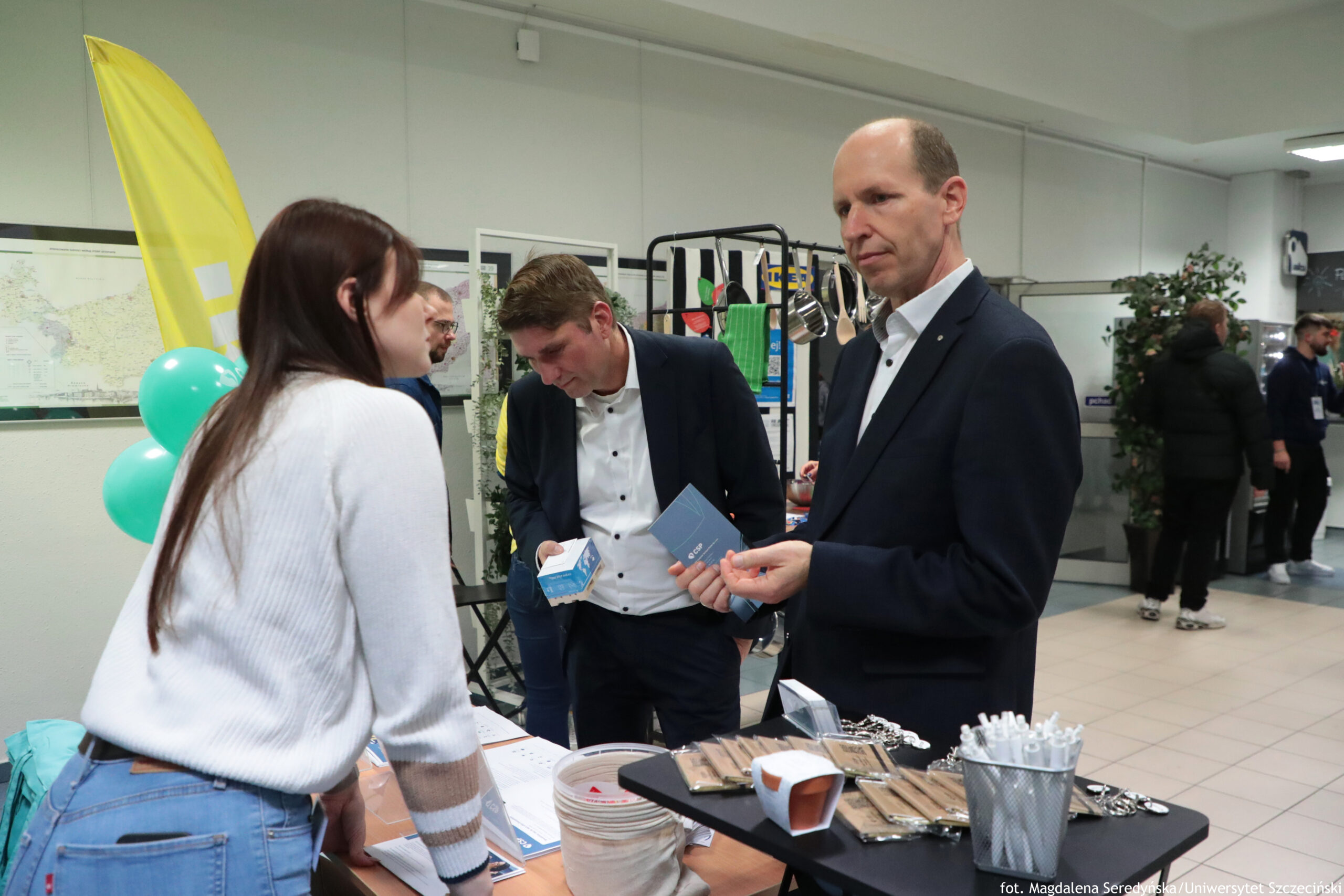Stoisko informacyjne w holu, z trzema osobami na pierwszym planie. Po lewej stoi młoda kobieta o jasnej skórze i długich, ciemnych włosach, w białym swetrze i niebieskich dżinsach; jest zwrócona bokiem i pochyla się nad stołem, rozmawiając z mężczyznami. Po prawej stoi wysoki mężczyzna o jasnej skórze, z bardzo krótkimi włosami i cofającą się linią włosów, w ciemnym garniturze i białej koszuli; trzyma w dłoniach niewielki niebieski kartonik/ulotkę. Obok niego, bliżej środka, drugi mężczyzna o jasnej skórze i brązowych włosach, w ciemnym garniturze i białej koszuli, pochyla głowę w dół i trzyma w ręku małe pudełko. Na stole leżą różne materiały i drobiazgi: ulotki, kilka drewnianych przedmiotów wyglądających, oraz metalowy pojemnik pełen białych długopisów. W tle widać żółtą flagę reklamową i zielone balony, a dalej kilka osób przechodzących oraz duże rośliny w donicach. W prawym dolnym rogu jest podpis: „fot. Magdalena Seredyńska / Uniwersytet Szczeciński”.
