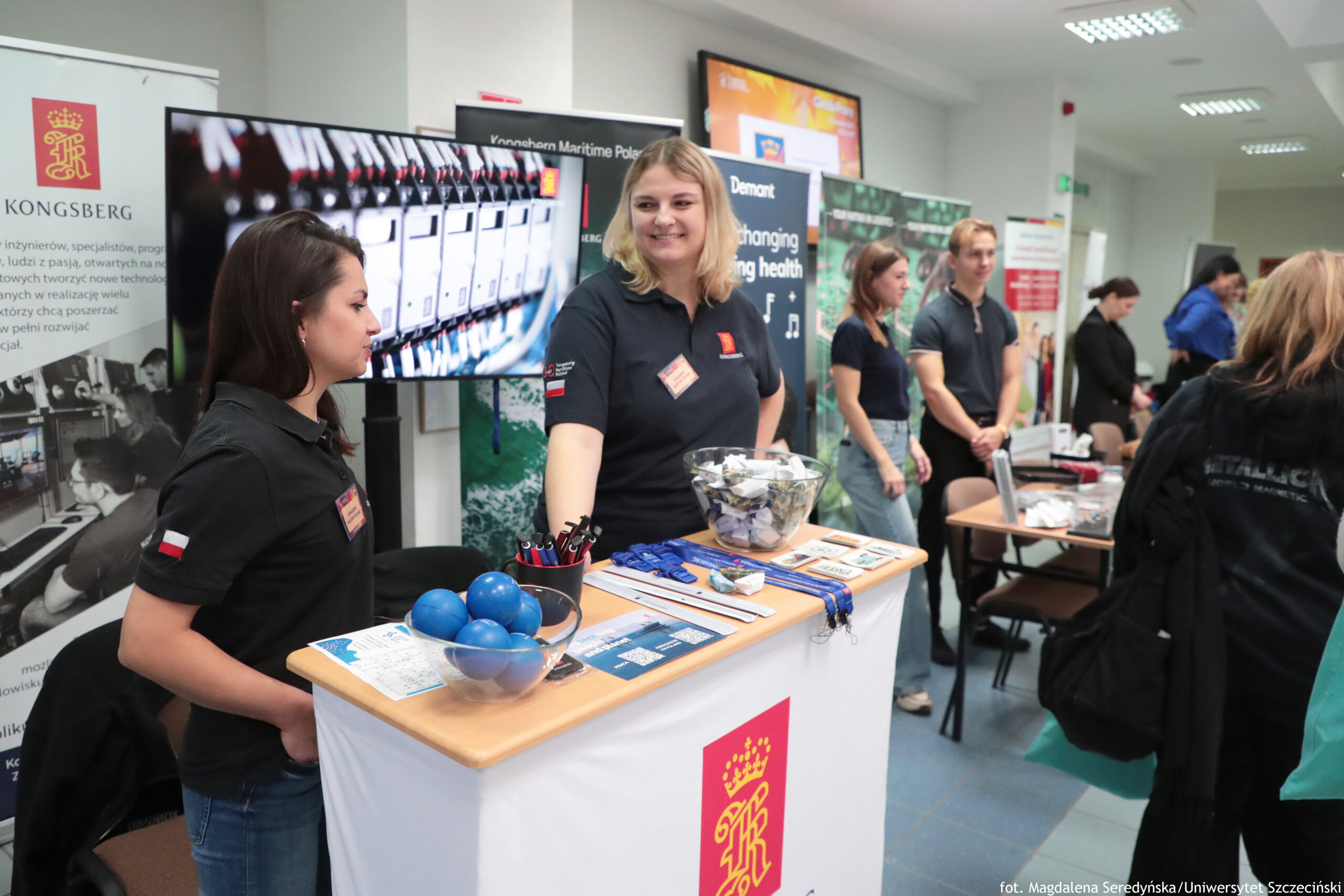 Stoisko informacyjne na targach lub wydarzeniu rekrutacyjnym w jasnej sali. Na pierwszym planie stoi lada z białą osłoną i czerwono-złotym logo. Na blacie leżą materiały promocyjne: smycze w kolorze niebieskim, długopisy w kubku, ulotki oraz dwie przezroczyste miski (jedna z niebieskimi piłeczkami, druga z karteczkami lub losami). Za ladą stoją dwie kobiety w czarnych koszulkach polo z identyfikatorami; jedna ma ciemne włosy i jest zwrócona bokiem, druga jest blondynką i uśmiecha się w stronę aparatu. W tle widać ekran z wyświetloną grafiką oraz kilka innych stoisk z osobami stojącymi i rozmawiającymi. Na jednym z banerów widoczny jest napis „KONGSBERG”.