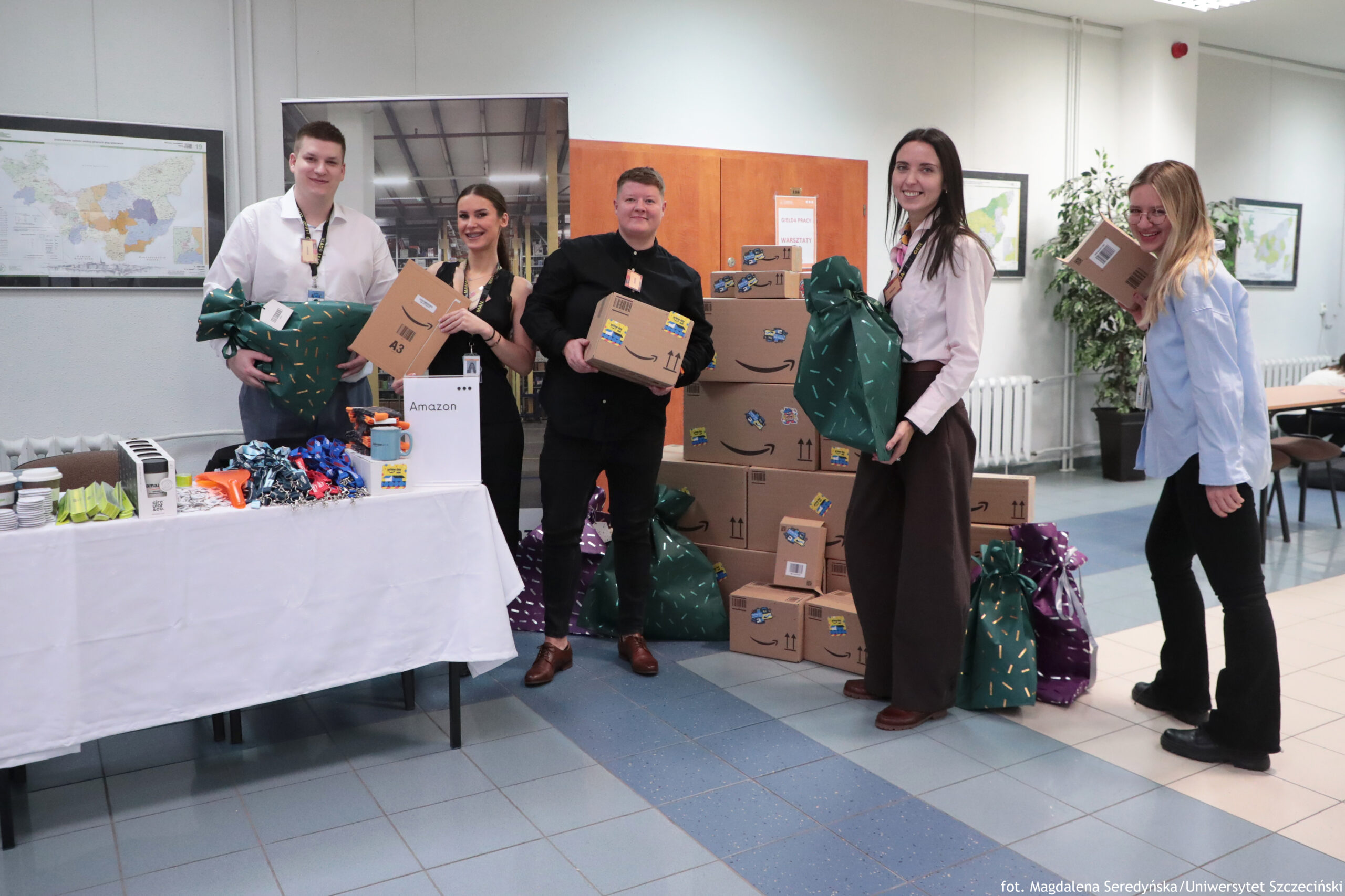 Grupa pięciu osób stojących w holu, obok stosu kartonów Amazon i dużych worków prezentowych. Wszyscy się uśmiechają i trzymają paczki lub worki. Po lewej stronie stoi mężczyzna w białej koszuli, trzyma duży zielony worek z wzorem. Obok niego kobieta w czarnej sukience trzyma płaski karton z naklejką. W środku stoi osoba w czarnym ubraniu, trzyma karton. Po prawej stoi wysoka kobieta w jasnej koszuli i długiej brązowej spódnicy, trzyma duży zielony worek. Najbardziej po prawej stoi kobieta w jasnoniebieskiej koszuli, trzyma małe pudełko. Z przodu po lewej jest stół przykryty białym obrusem, na nim leżą drobne przedmioty i kolorowe wstążki; stoi też biały stojak z napisem „Amazon”. W tle widać ściany z mapami w ramach, roślinę w donicy i część biurek. W prawym dolnym rogu jest mały podpis: „fot. Magdalena Seredyńska / Uniwersytet Szczeciński”.