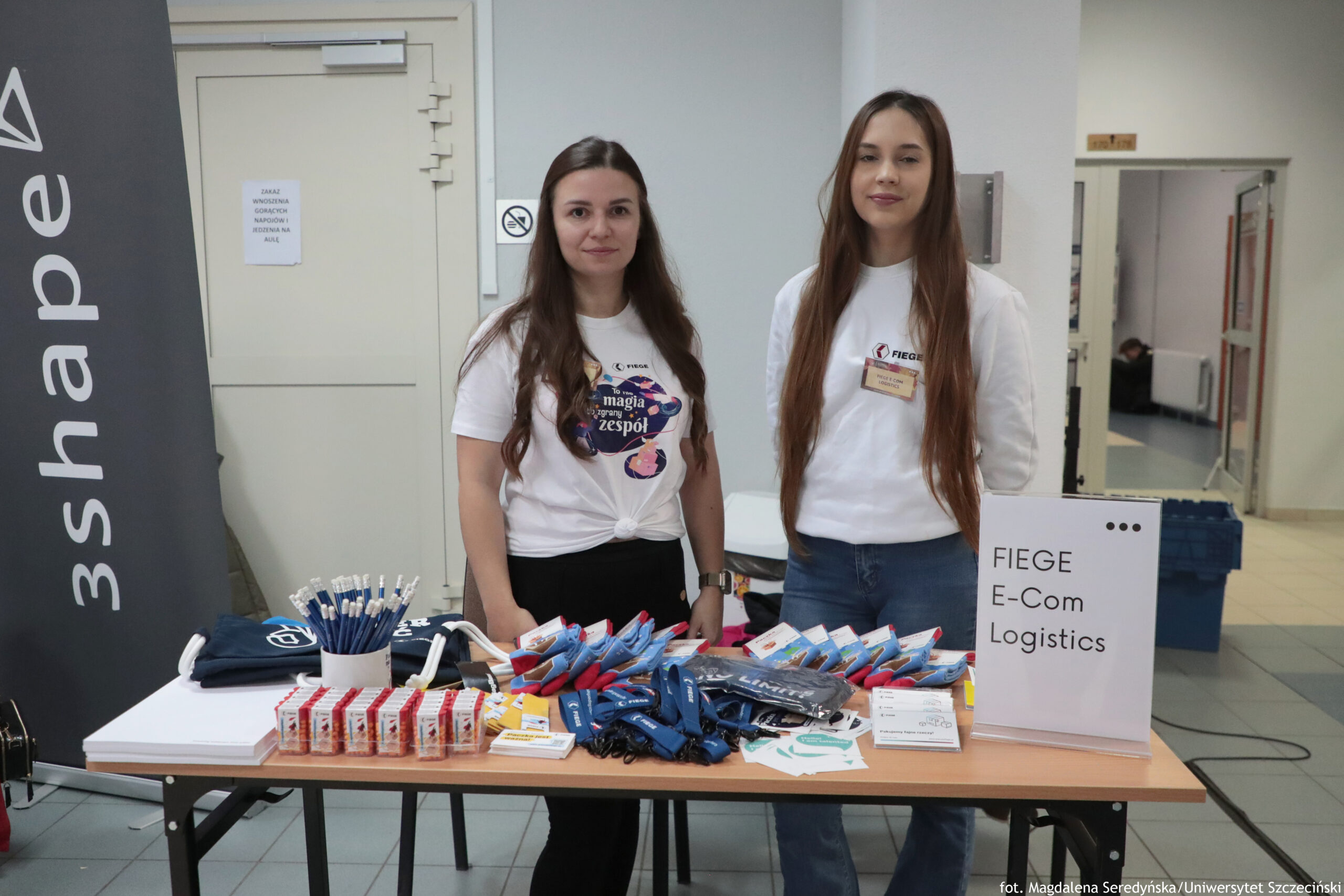 Stoisko informacyjne z dwoma kobietami stojącymi za stołem z materiałami promocyjnymi w budynku. Kobieta po lewej ma długie, ciemne włosy i jasną skórę. Ma na sobie białą koszulkę z dużą, kolorową grafiką i napisem „magia zgrany zespół”; koszulka jest związana na dole. Patrzy w stronę aparatu z neutralnym wyrazem twarzy. Kobieta po prawej ma bardzo długie, proste, jasnobrązowe włosy i jasną skórę. Ma na sobie białą koszulkę z logo „FIEGE” i identyfikator na smyczy. Stoi z lekko uśmiechniętą twarzą. Na stole leżą i stoją: kubek z wieloma długopisami, stosy małych czerwono-białych pudełek, rozłożone smycze i breloki w kolorze niebieskim, kilka ulotek i kart oraz ciemne materiałowe gadżety (np. torby lub koszulki). Po prawej stronie stołu stoi biała tabliczka z napisem: „FIEGE E-Com Logistics”. Po lewej stronie kadru widać wysoki, ciemny baner z dużym, pionowym napisem „3shape”. W tle jest jasny korytarz lub hol, drzwi oraz kartka na drzwiach (tekst jest zbyt mały, by odczytać w całości). Na ścianie obok jest znak zakazu palenia. W prawym dolnym rogu zdjęcia widnieje podpis: „fot. Magdalena Seredyńska/Uniwersytet Szczeciński”.