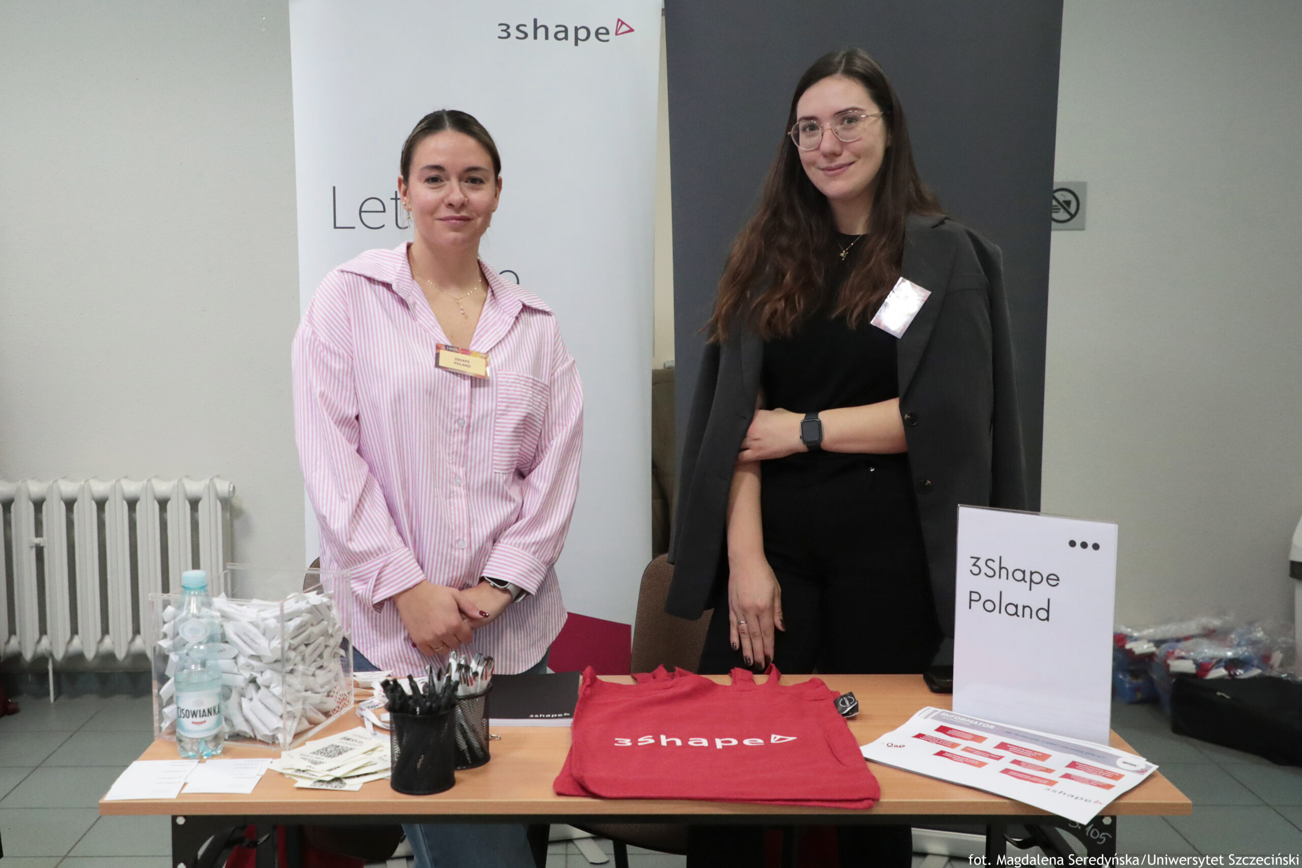 Dwie kobiety stojące za stołem na stoisku promocyjnym z logo „3shape”. Po lewej kobieta o jasnej karnacji, z ciemnymi włosami związanymi z tyłu, w różowo-białej koszuli w paski i z identyfikatorem; po prawej kobieta o jasnej karnacji, z długimi ciemnymi włosami, w okularach, ubrana na czarno, z ciemną marynarką narzuconą na ramiona i z identyfikatorem. Na stole leżą materiały promocyjne: czerwona koszulka z napisem „3shape”, stojak z kartką „3Shape Poland”, ulotki i kartki, czarny pojemnik z długopisami, przezroczyste pudełko wypełnione złożonymi papierkami oraz butelka wody. W tle widać roll-up z napisem „3shape”, grzejnik i ścianę. W prawym dolnym rogu jest podpis: „fot. Magdalena Seryńska / Uniwersytet Szczeciński”.