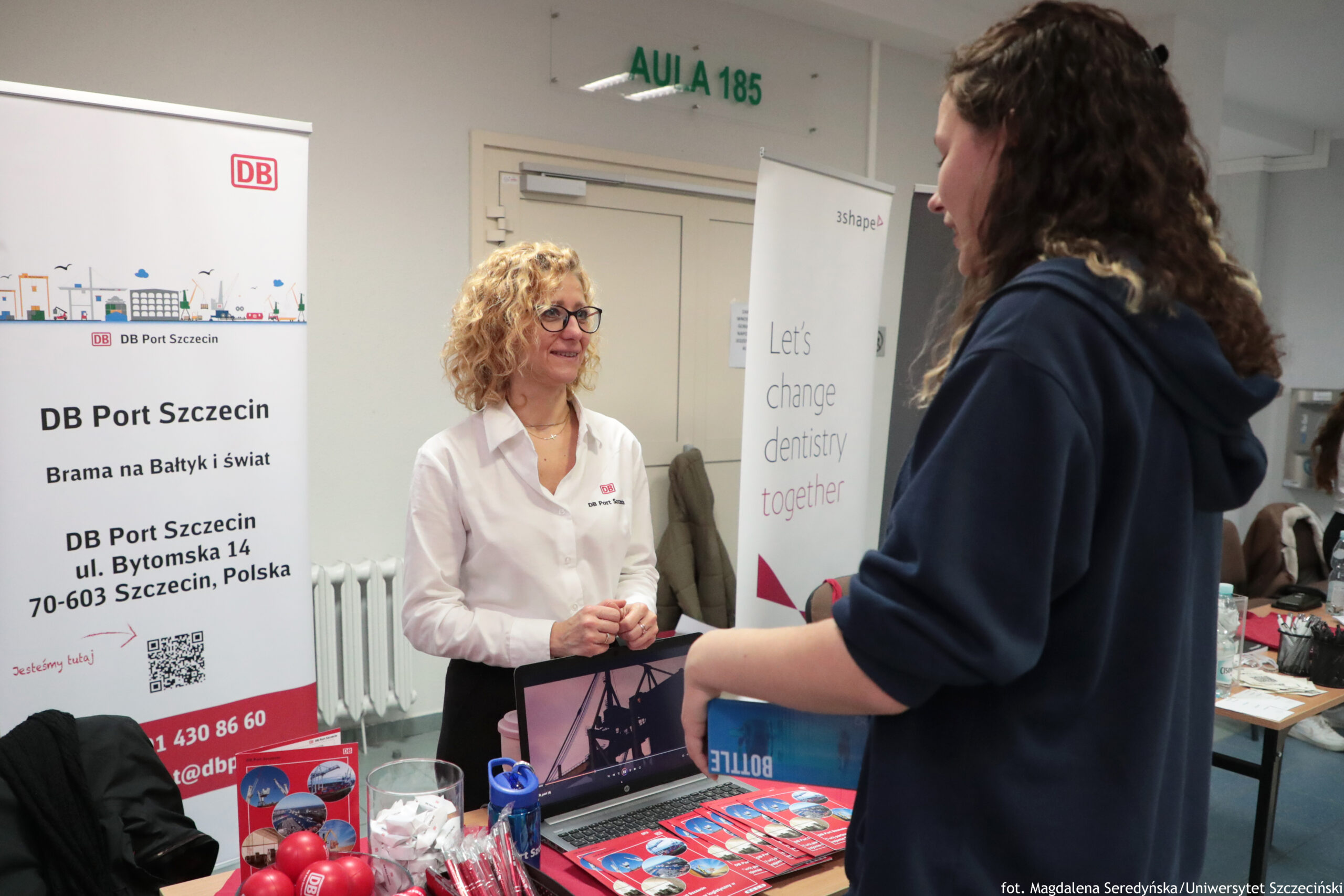 Stoisko informacyjne na uczelni z dwiema osobami rozmawiającymi przy stole. Kobieta w średnim wieku z jasnymi, kręconymi włosami i okularami ma na sobie białą koszulę z logo „DB” i uśmiecha się, stojąc za stołem. Naprzeciwko stoi młoda osoba z długimi, falowanymi brązowymi włosami w granatowej bluzie. Po lewej stronie widać roll-up z napisem: „DB Port Szczecin”, „Brama na Bałtyk i świat”, „DB Port Szczecin ul. Bytomska 14 70-603 Szczecin, Polska”. Na stole leżą czerwone ulotki, długopisy, małe gadżety i otwarty laptop. W tle nad drzwiami znajduje się zielony napis „AULA 185”. Po prawej stronie stoi drugi roll-up z tekstem „Let’s change dentistry together” i logo „3Shape”. W prawym dolnym rogu jest podpis: „fot. Magdalena Seredyńska / Uniwersytet Szczeciński”.