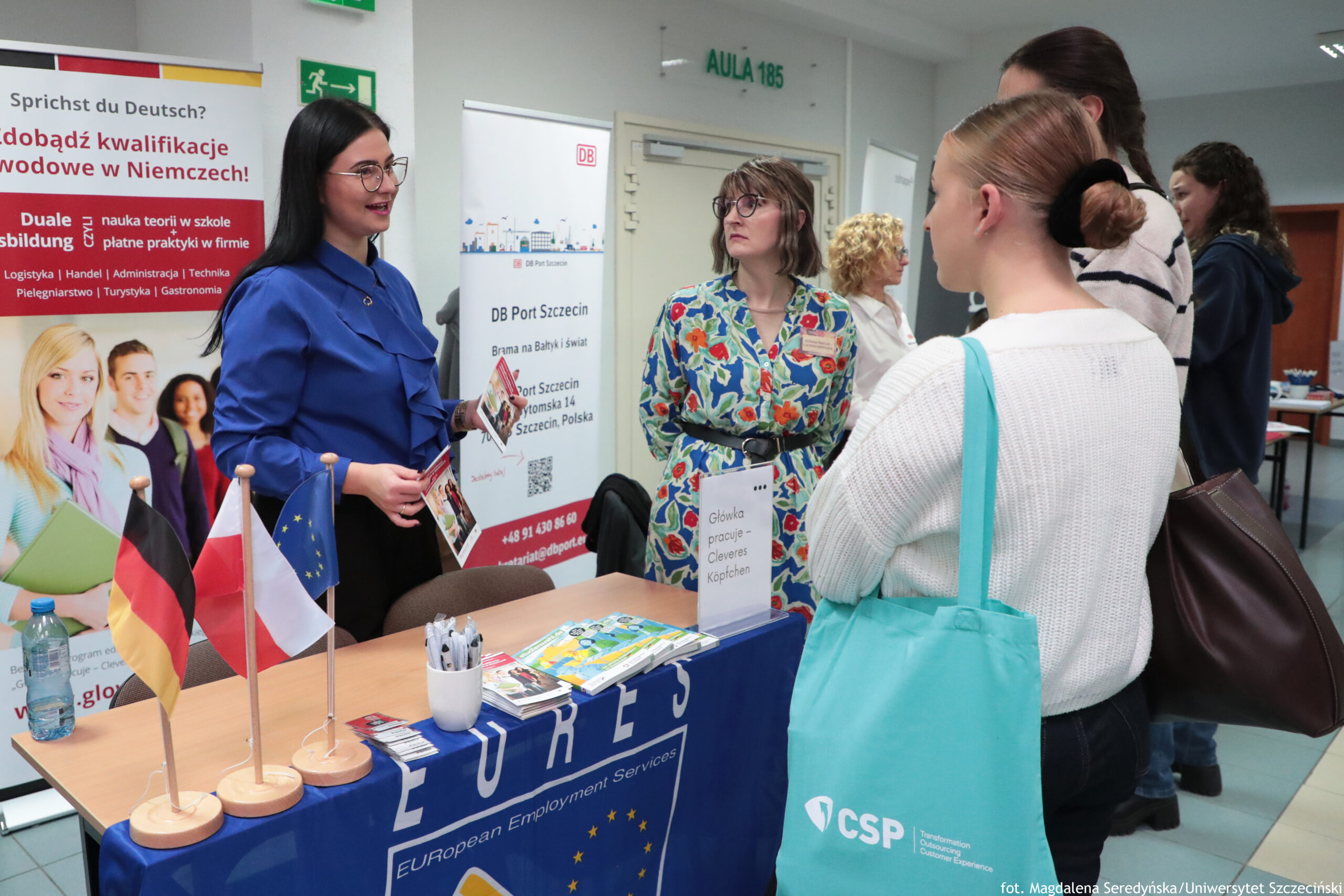 Stół informacyjny na targach pracy „EURES European Employment Services” i małymi flagami Niemiec, Polski oraz Unii Europejskiej na drewnianych podstawkach. Na blacie leżą ulotki i katalogi, stoi kubek z długopisami oraz butelka wody; na stojaku widoczna kartka z napisem „Główka pracuje – Cleveres Köpfchen”. Przy stoisku stoi młoda kobieta o jasnej skórze, z długimi czarnymi włosami i okularami, w niebieskiej koszuli; trzyma ulotki i rozmawia z osobami po drugiej stronie stołu. Naprzeciwko stoi kobieta o jasnej skórze z blond włosami upiętymi w kok, w białym swetrze, z turkusową torbą na ramieniu. Obok niej widać jeszcze kilka osób stojących w kolejce. W tle znajdują się roll-upy reklamowe dotyczące pracy i kształcenia w Niemczech, w tym „DB Port Szczecin”, oraz drzwi z zielonym napisem „AULA 105”. W prawym dolnym rogu widnieje podpis: „fot. Magdalena Seredyńska/Uniwersytet Szczeciński”.