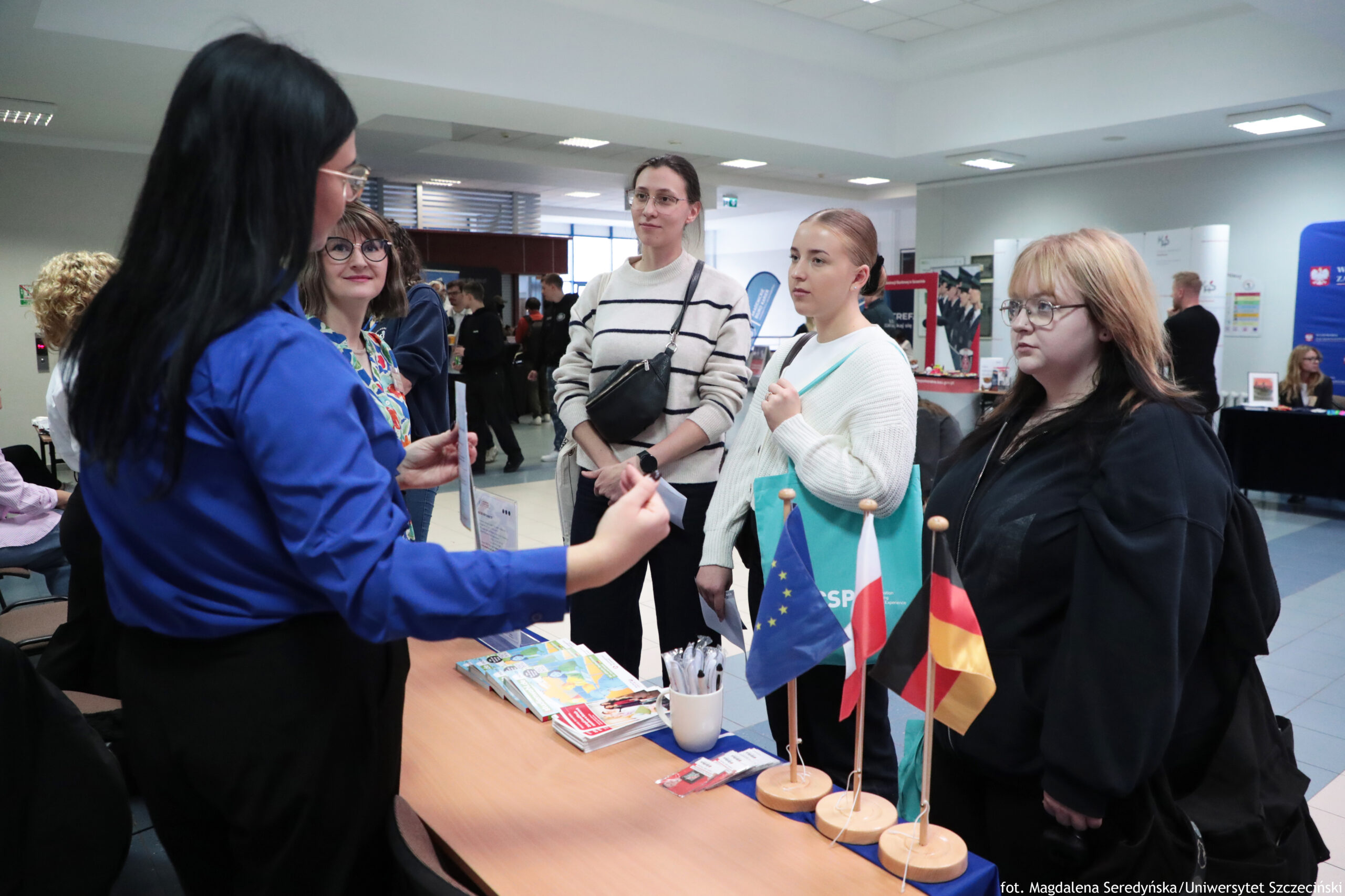 Stół informacyjny w holu wewnątrz budynku, z małymi flagami Unii Europejskiej, Polski i Niemiec ustawionymi na drewnianych podstawkach. Na blacie leżą ulotki i broszury, a także kubek z długopisami. Po lewej stronie stoi kobieta o długich, czarnych włosach w niebieskiej koszuli, zwrócona bokiem i gestykulująca podczas rozmowy. Naprzeciwko niej, po drugiej stronie stołu, stoją trzy kobiety: jedna w okularach i kolorowej koszuli, druga wyższa w jasnym swetrze w czarne paski z czarną torebką przewieszoną przez ramię, oraz trzecia w jasnym swetrze, z włosami związanymi z tyłu, trzymająca turkusową torbę. Po prawej stronie stoi jeszcze jedna kobieta w okularach, z jasnobrązowymi włosami, ubrana na czarno, skierowana w stronę stołu. W tle widać inne osoby oraz stoiska i banery. W prawym dolnym rogu jest podpis: „fot. Magdalena Seredyńska/Uniwersytet Szczeciński”.