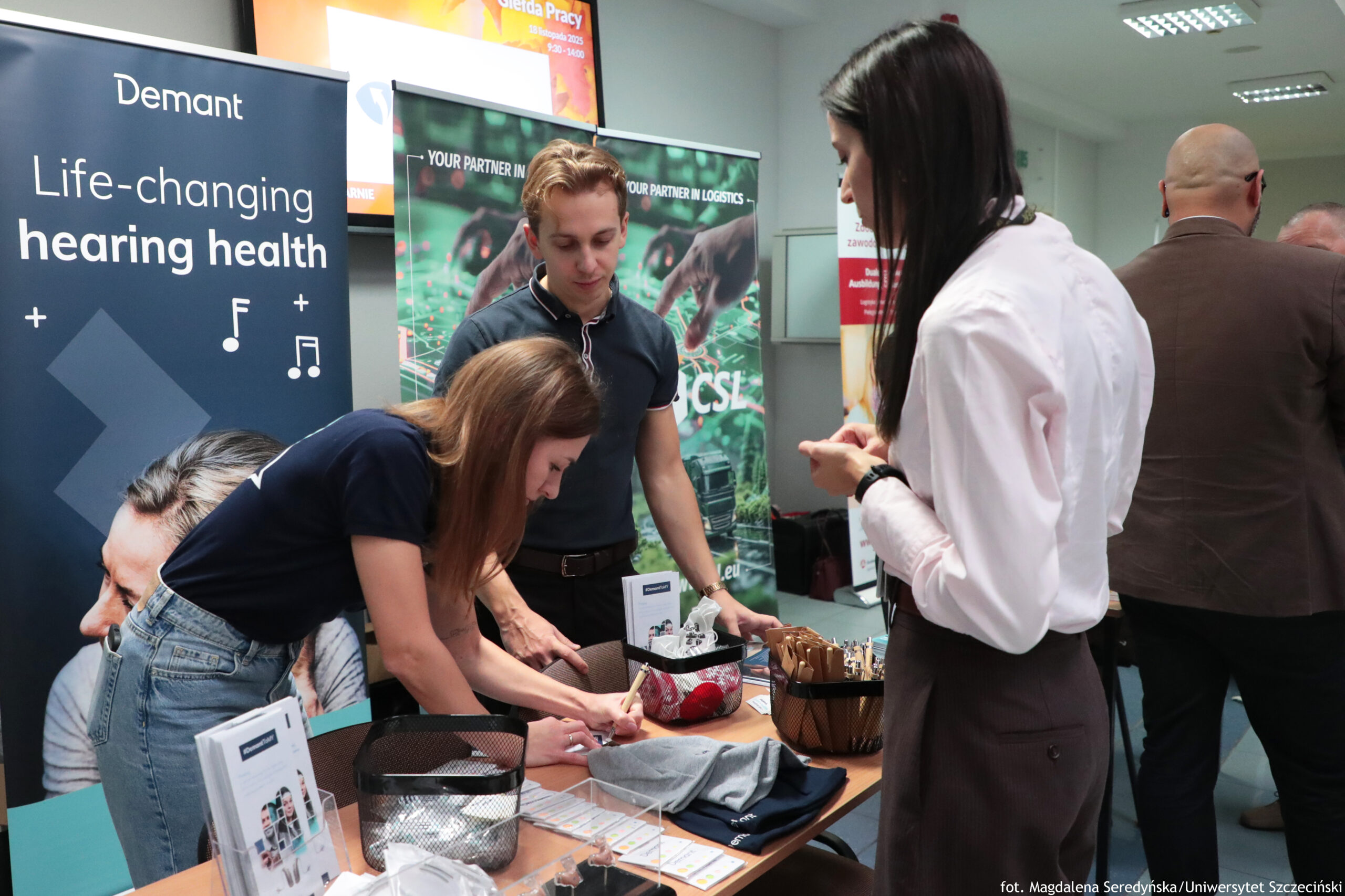 Stoisko informacyjne na wydarzeniu w pomieszczeniu, z trzema osobami stojącymi przy stole i banerami firmowymi w tle. Na stole leżą ulotki, formularze do podpisu, długopisy, wizytówki, siatkowy koszyk, pudełko z drobnymi upominkami oraz złożone ubrania lub tekstylia w kolorach szarym i granatowym. Po lewej stronie stoi baner z napisem: “Demant” oraz “Life-changing hearing health”, z ikonami nut i symbolami “+”. Obok w tle widać drugi baner z napisem u góry: “YOUR PARTNER IN LOGISTICS”, z fotografią dłoni nad zielonym tłem i częściowo widocznym logo/napisem (nie w całości czytelny). W prawym dolnym rogu zdjęcia znajduje się podpis: “fot. Magdalena Seredyńska/Uniwersytet Szczeciński”. Przy stole kobieta z długimi, rudobrązowymi włosami pochyla się i pisze na formularzu. Za nią stoi mężczyzna o jasnych włosach w ciemnej koszulce polo, patrzący w dół na stół. Po prawej stronie stoi kobieta o długich, ciemnych włosach w białej koszuli i ciemnych spodniach. W tle po prawej widać jeszcze dwie osoby odwrócone tyłem.