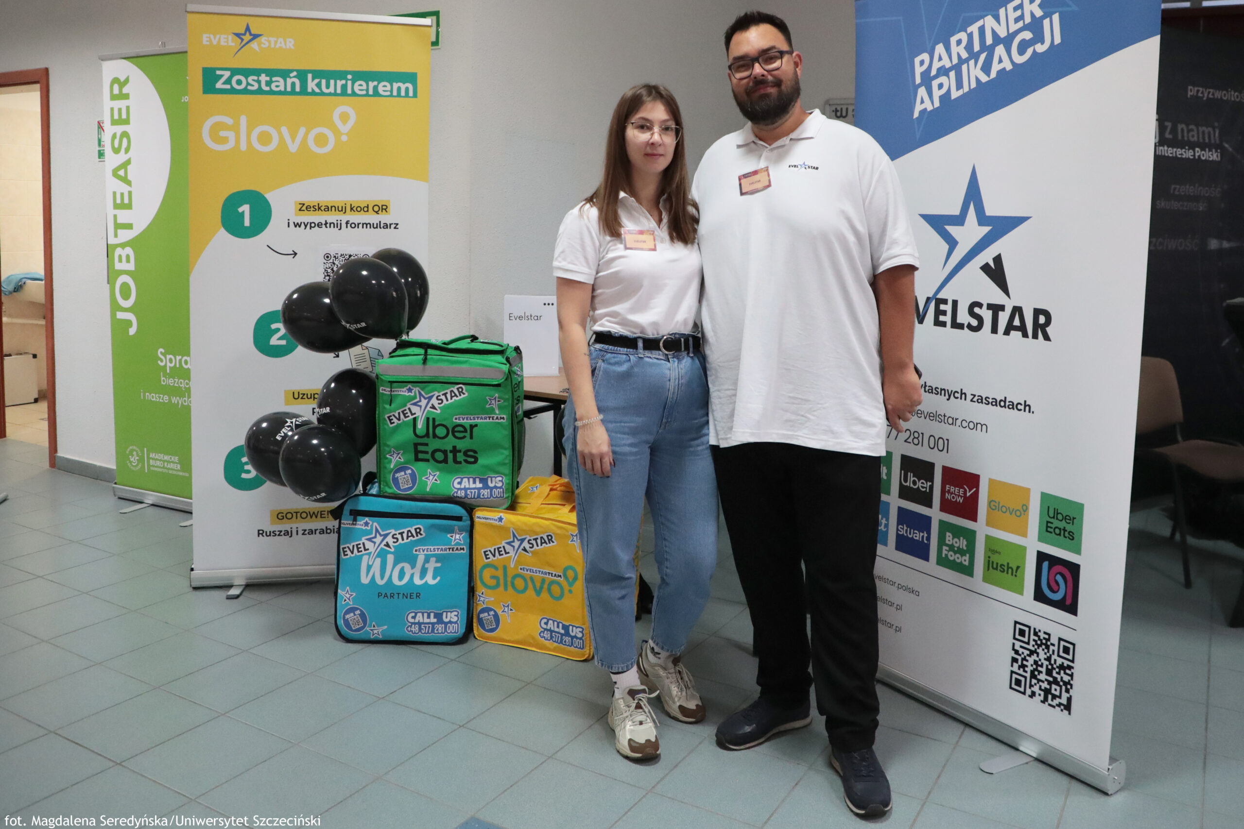 Dwóch dorosłych ludzi stojących w holu między roll-upami reklamowymi, obok kolorowych toreb/plecaków kurierskich i czarnych balonów. Osoby mają jasną karnację i ubrane są w białe koszulki polo z identyfikatorami; kobieta ma długie proste brązowe włosy, okulary, jasnoniebieskie jeansy i sportowe buty, a mężczyzna ma krótkie ciemne włosy, brodę i wąsy, okulary, ciemne spodnie i ciemne buty.