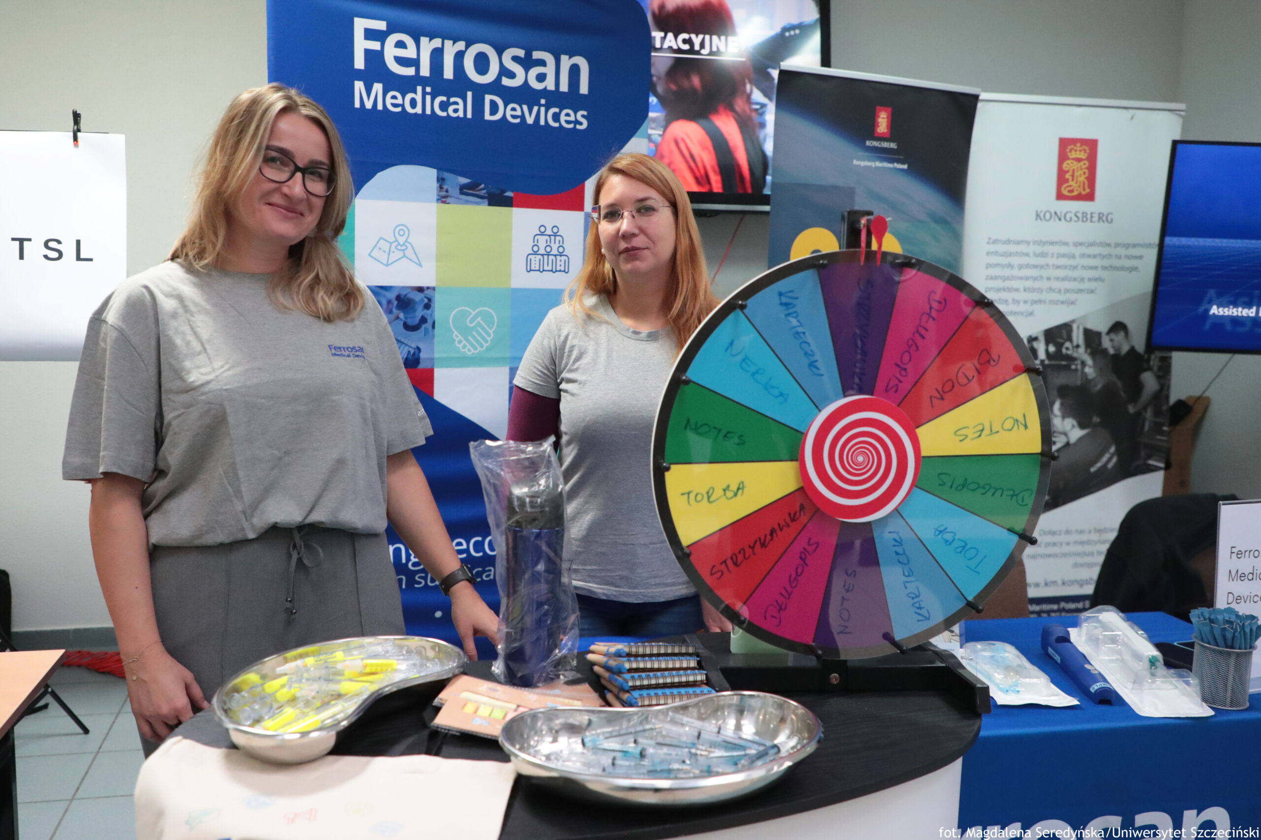 Stoisko promocyjne z dwoma kobietami stojącymi za ladą oraz dużym kolorowym kołem losującym na blacie. Kobiety mają jasną karnację; obie są w szarych koszulkach z logo. Kobieta po lewej ma długie blond włosy, okulary i uśmiecha się; ma też szare spodnie. Kobieta po prawej ma proste, rudawe włosy do ramion i okulary. Na pierwszym planie, na ladzie, stoi duże koło podzielone na wielokolorowe pola z napisami (część z nich jest czytelna): „NOTES”, „TORBA”, „KUBECZKI”, „STERYLNA”, „DŁUGOPIS”, „BIDON”. W środku koła jest czerwono-biała spirala. Na blacie leżą także metalowe tacki z drobnymi przedmiotami (wyglądają jak gadżety), kilka stosów notesów, zapakowane długopisy lub inne drobiazgi w folii oraz niebieskie opaski/smycze. W tle widać roll-upy i banery, m.in. duży napis „Ferrosan Medical Devices” oraz logo i napis „KONGSBERG”. Na dole po prawej jest mały napis kredytowy: „fot. Magdalena Seredyńska/Uniwersytet Szczeciński”.