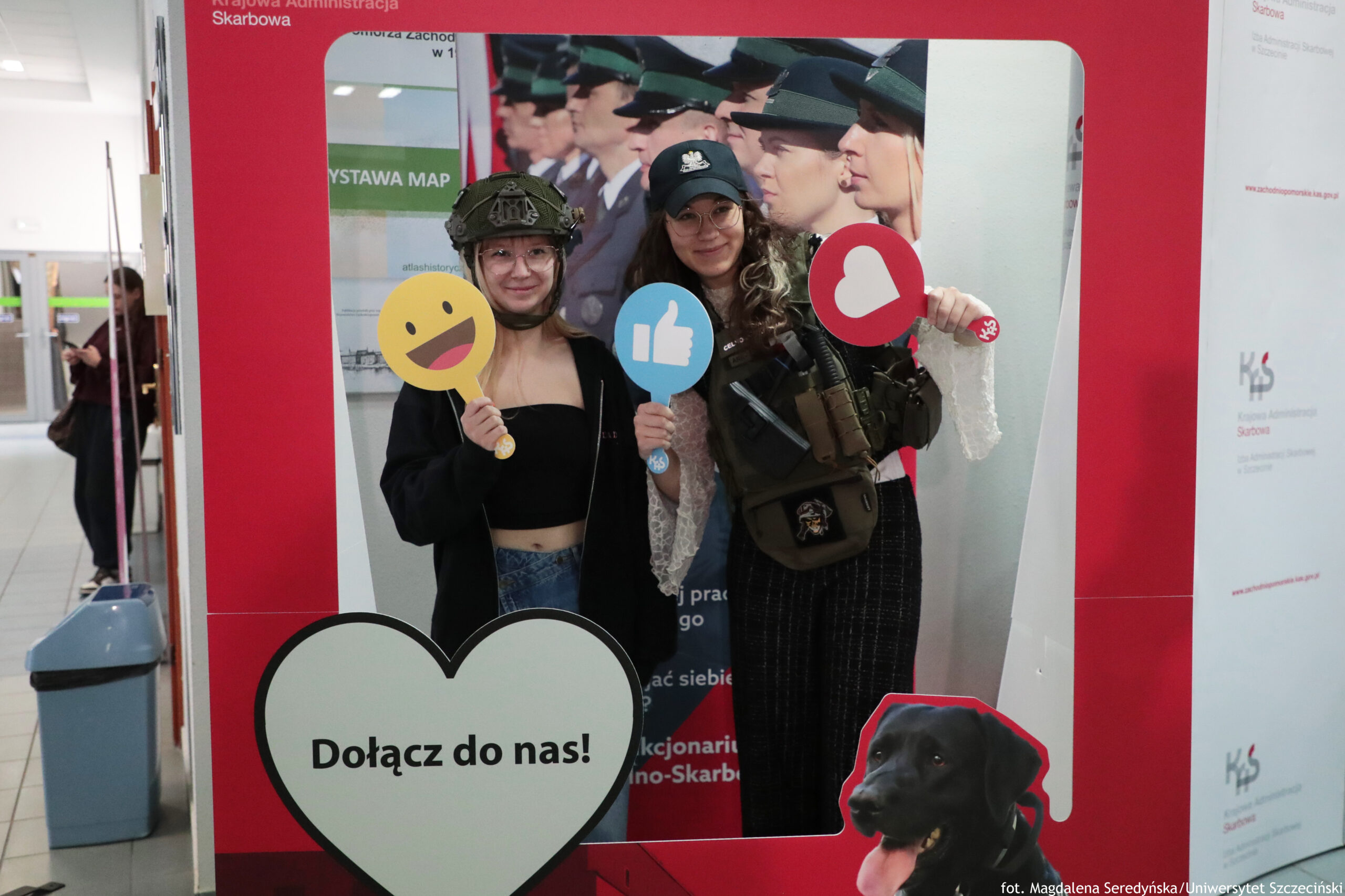 Czerwono-biała ścianka promocyjna z dużą ramką do zdjęć i napisem „Krajowa Administracja Skarbowa” u góry. W otworze ramki stoją dwie młode kobiety o jasnej karnacji, uśmiechnięte, w okularach. Kobieta po lewej ma na głowie wojskowy hełm, czarny top odsłaniający brzuch i trzyma okrągłą tabliczkę z żółtą buźką–emotikoną. Kobieta po prawej ma czapkę z daszkiem z białym godłem, kamizelkę taktyczną i trzyma dwie tabliczki: niebieską z kciukiem w górę oraz czerwoną z białym sercem. Na dole ramki jest duże białe serce z napisem „Dołącz do nas!”. W prawym dolnym rogu grafika czarnego psa z wysuniętym językiem. W tle, na plakacie za nimi, widoczny jest rząd umundurowanych osób w czapkach. W prawym dolnym rogu zdjęcia jest podpis: „fot. Magdalena Seredyńska/Uniwersytet Szczeciński”.