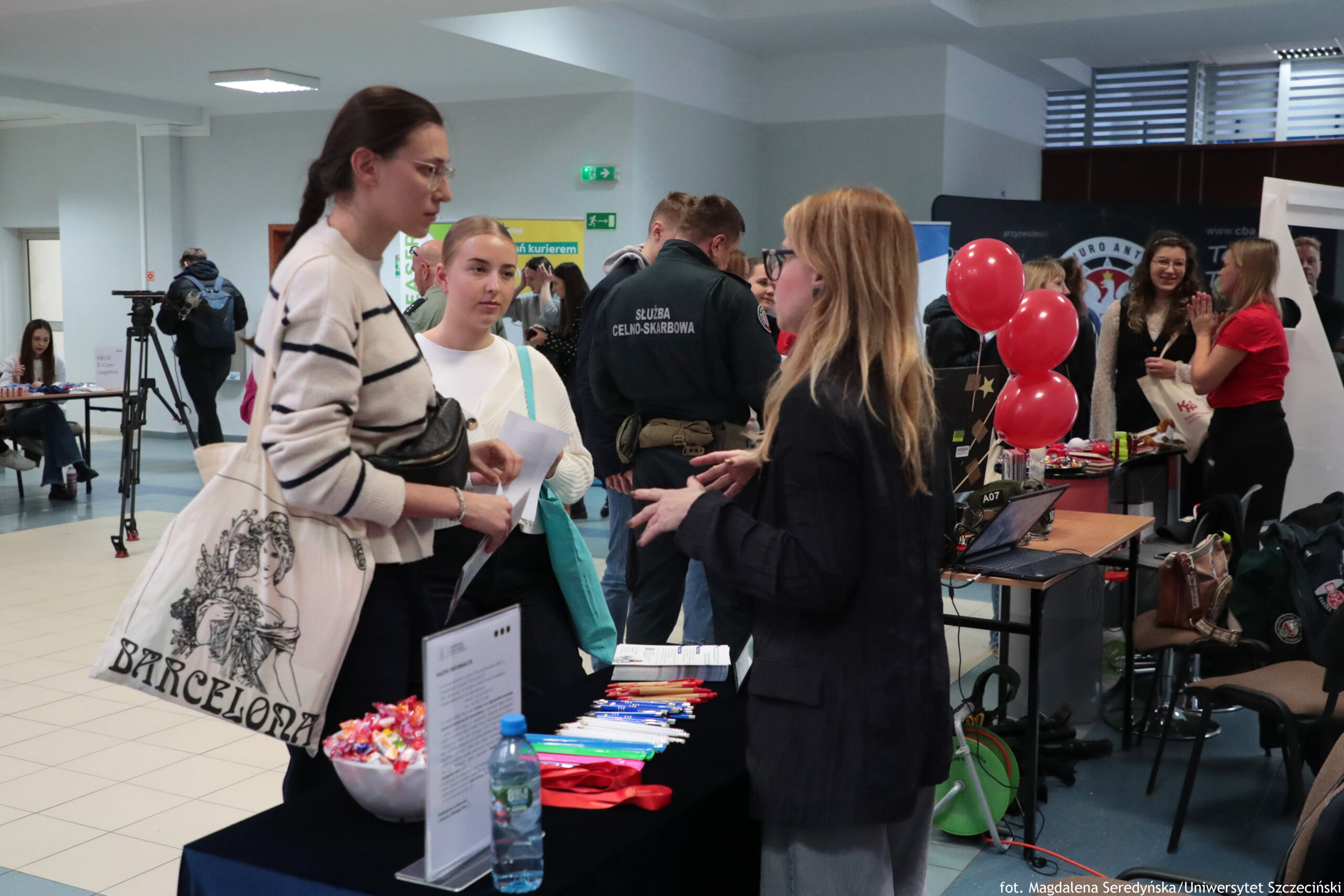 Stoisko informacyjne na holu, przy którym dwie kobiety rozmawiają; na stole leżą długopisy, smycze i materiały promocyjne, stoi też butelka wody i misa z cukierkami. W tle widać tłum osób przy kolejnych stoiskach, czerwone balony, laptop na biurku oraz osobę w mundurze z napisem „SŁUŻBA CELNO-SKARBOWA”. Po lewej stronie znajduje się statyw z kamerą oraz stoliki z siedzącymi osobami. Na torbie materiałowej jednej z kobiet widoczny jest napis „BARCELONA”. W prawym dolnym rogu zdjęcia jest podpis: „fot. Magdalena Seredyńska/Uniwersytet Szczeciński”.