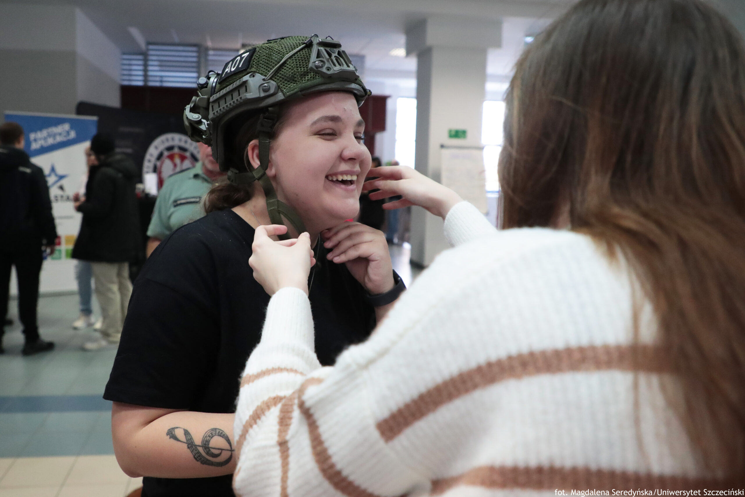 Uśmiechnięta kobieta w zielonym hełmie ochronnym z paskiem pod brodą. Druga osoba stoi przodem do niej i zapina lub poprawia pasek przy hełmie. Kobieta ma jasną skórę, ciemne włosy związane z tyłu, czarną koszulkę i widoczny tatuaż na przedramieniu (motyw nut i klucza wiolinowego). Zdjęcie w holu podczas wydarzenia, w tle są rozmyte osoby i stojące tablice/plakaty. W prawym dolnym rogu widoczny napis: „fot. Magdalena Seredyńska/Uniwersytet Szczeciński”.