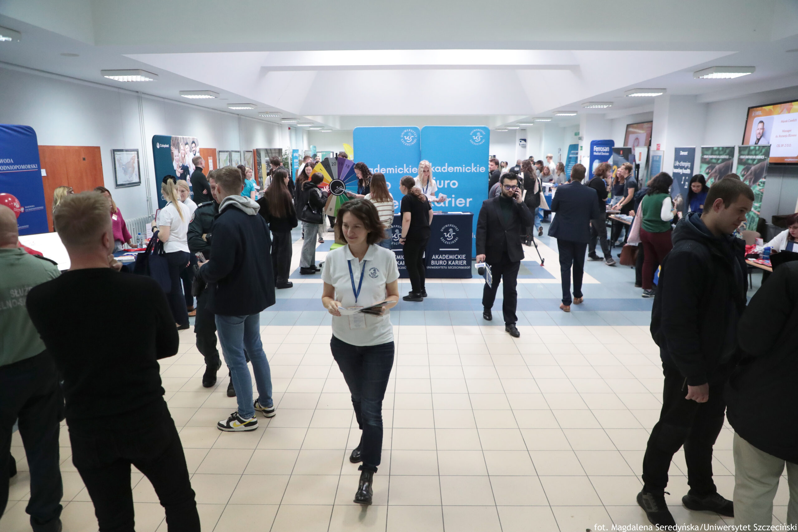 Zatłoczony hol wewnątrz budynku, z wieloma stoiskami i banerami. W kadrze jest kilkadziesiąt osób stojących w grupkach i rozmawiających; na środku idzie kobieta w białej koszulce polo z identyfikatorem na smyczy i trzyma ulotki lub kartki. W tle pośrodku stoją duże niebieskie ścianki z napisem „Akademickie Biuro Karier”. Obok widać kolorowe koło/ruletkę promocyjną. Po bokach ustawione są stoiska z plakatami i roll-upami; część osób stoi przy stołach i przegląda materiały. Pomieszczenie jest jasne, z białym sufitem i wieloma prostokątnymi lampami, a podłoga jest z jasnych płytek. W prawym górnym rogu jest ekran z wyświetlaną reklamą/ogłoszeniem, ale tekst jest za mały, żeby go dokładnie odczytać. W prawym dolnym rogu widać podpis: „fot. Magdalena Seredyńska / Uniwersytet Szczeciński”.