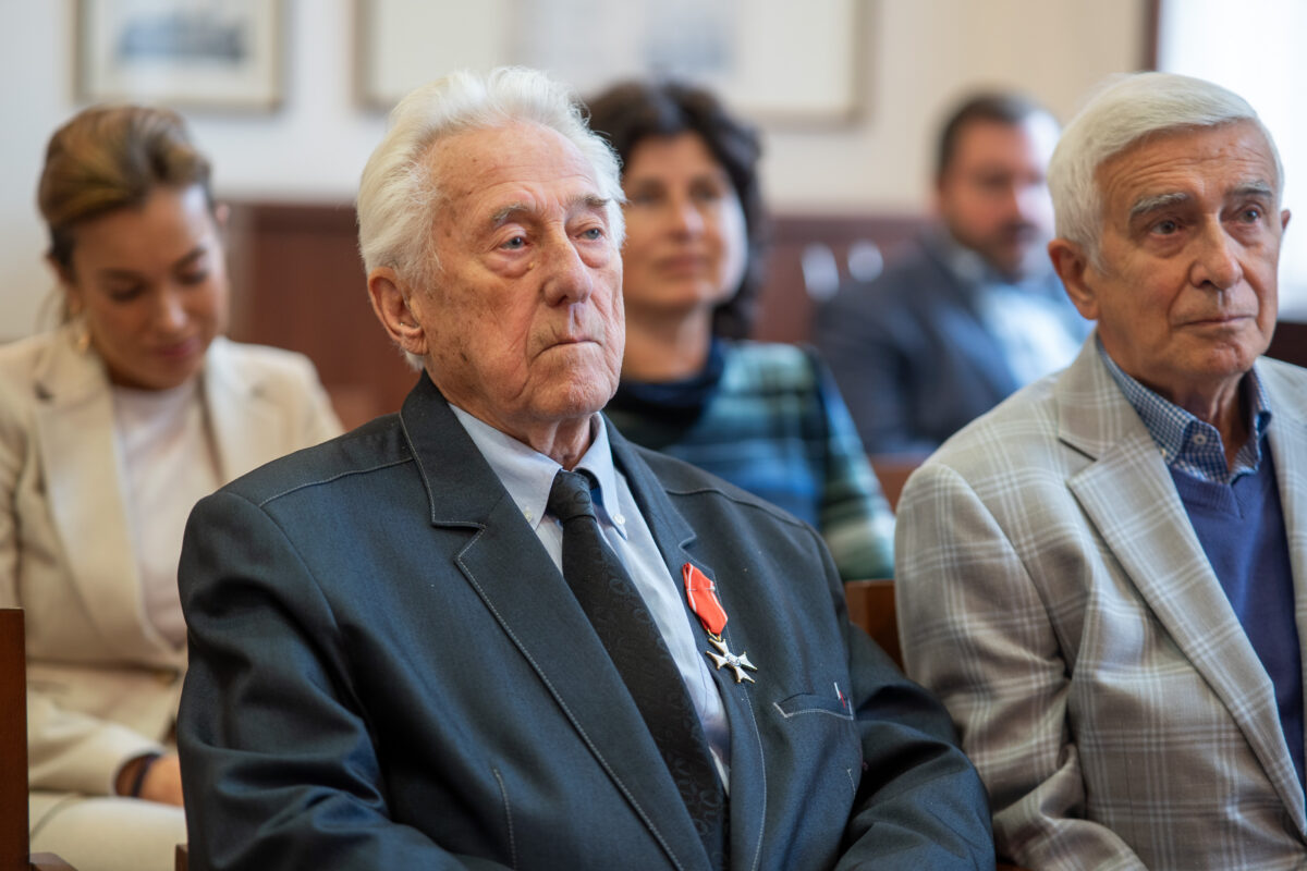 Na pierwszym planie dwóch mężczyzn, w tym prof. Tadeusz Białecki odznaczony Krzyżem Kawalerskim Orderu Odrodzenia Polski (ujęcie do pasa). W tle ludzie i ściana archiwalnymi czarno-białymi fotografiami.