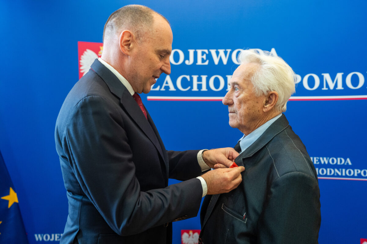 Wojewoda Zachodniopomorski – dr Adam Rudawski (po lewej) wpina w marynarkę odznaczenie prof. Tadeuszowi Białeckiemu. W tle niebieska ścianka z godłem Polski i białym napisem: Wojewoda Zachodniopomorski.