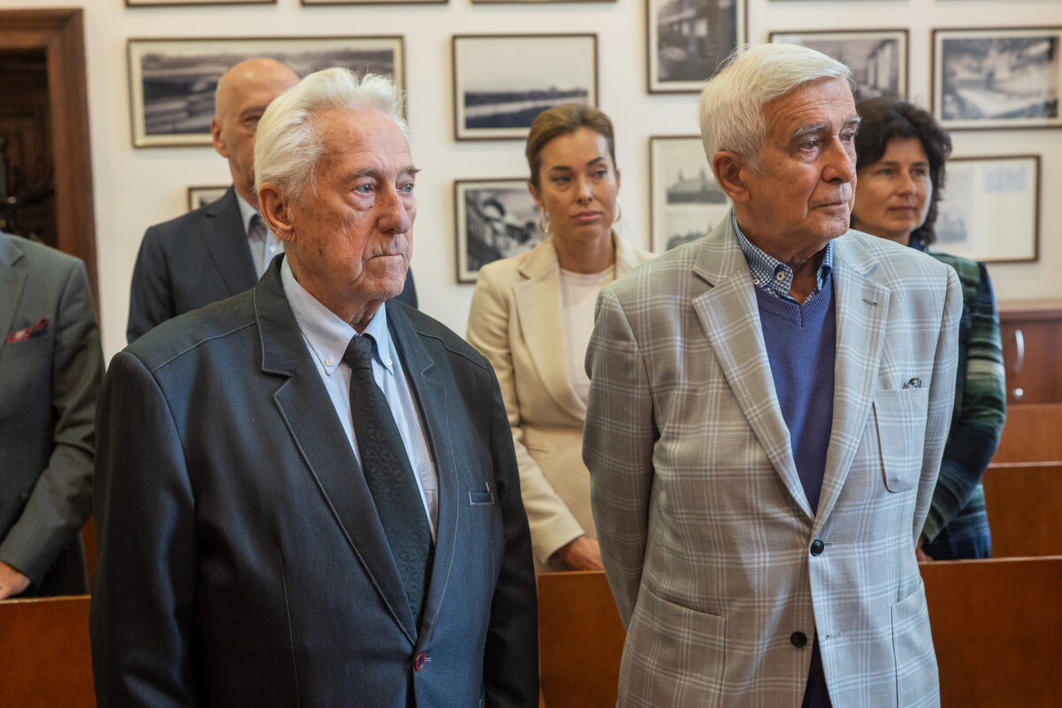 Na pierwszym planie dwóch mężczyzn, w tym prof. Tadeusz Białecki odznaczony Krzyżem Kawalerskim Orderu Odrodzenia Polski (ujęcie do pasa). W tle ludzie i ściana archiwalnymi czarno-białymi fotografiami.