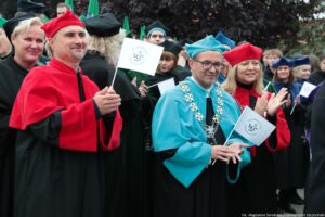 Spacerek na uniwerek. Ujęcie przodem. Idący przedstawiciele Uniwersytetu Szczecińskiego ubrani w czarne togi z zielonymi, czerwonymi i niebieskimi elementami, trzymający białe papierowe flagi. W tle drzewa.