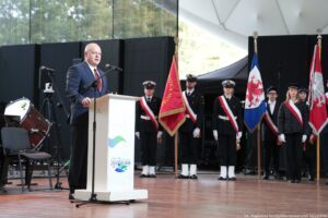 Inauguracja roku akademickiego. Scena. Wiceminister nauki i szkolnictwa wyższego stojący za białą mównicą. Przed nim mikrofon. W tle po prawej poczty sztandarowe.