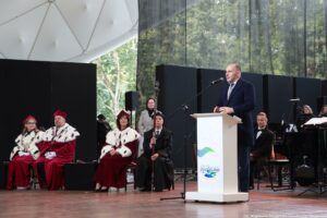 Inauguracja roku akademickiego. Scena. Wojewoda zachodniopomorski stojący za białą mównicą. Przed nim mikrofon. W tle po prawej instrumenty i chór. Po lewej przedstawiciele uczelni, w tym troje rektorów w czerwonych togach z gronostajami.