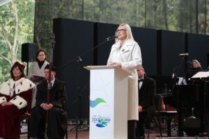 Inauguracja roku akademickiego. Scena. Wicemarszałkini województwa zachodniopomorskiego stojąca za białą mównicą. Przed nią mikrofon. W tle po prawej instrumenty i chór. Po lewej przedstawiciele uczelni – rektorka, pedel i rzeczniczka prasowa.