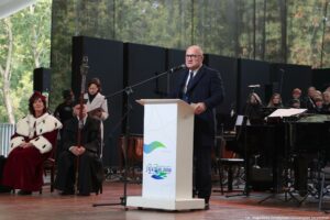 Inauguracja roku akademickiego. Scena. Prezydent Szczecina stojący za białą mównicą. Przed nim mikrofon. W tle po prawej instrumenty i chór. Po lewej przedstawiciele uczelni – rektorka, pedel i rzeczniczka prasowa.