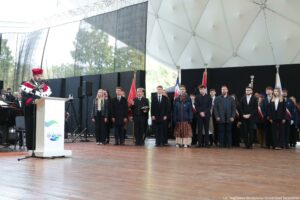 Inauguracja roku akademickiego. Scena. Po lewej przedstawiciel uczelni w uroczystej todze z gronostajami za białą mównicą. Po prawej immatrykulowani studenci, za nimi poczty sztandarowe.