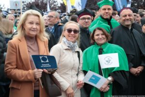 Spacerek na uniwerek. Przedstawiciele Uniwersytetu Szczecińskiego ubrani w czarne togi z zielonymi i bordowymi elementami, trzymający białe papierowe flagi z logiem uczelni, Spacerku na uniwerek i Akademickiego Szczecina. W tle inni uczestnicy wydarzenia.