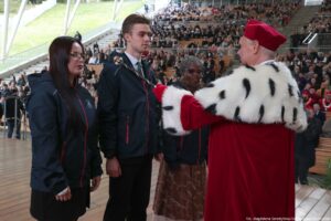 Inauguracja roku akademickiego – immatrykulacja. Ujęcie bokiem. Scena. Po prawej rektor Uniwersytetu Szczecińskiego z berłem, ubrany w czerwoną togę z gronostajami. Po lewej troje studentów (dwie kobiety, jeden mężczyzna – w środku) w granatowych kurtkach z białym logiem uczelni na piersi. W tle widownia Teatru Letniego.