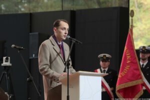 Scena. Mężczyzna stojący za białą mównicą. Za nim i przed nim mikrofon. W tle po prawej dwie osoby trzymające sztandar.