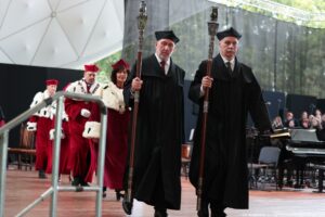 Scena. Z przodu dwóch pedli w czarnych togach. Za nimi schodzący ze sceny rektorzy szczecińskich uczelni w togach z czerwonymi elementami i gronostajami. W tle po prawej fortepiany i chór.