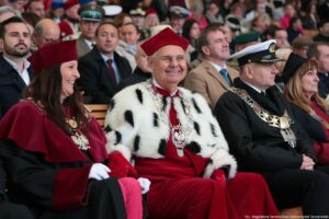 Ujęcie bokiem. Inauguracja roku akademickiego. Siedząca widownia. W pierwszym rzędzie władze szczecińskich uczelni – w środku rektor Uniwersytetu Szczecińskiego w todze z czerwonymi elementami i gronostajami. W tle pozostali uczestnicy wydarzenia.
