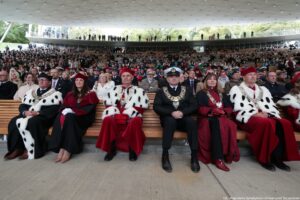 Ujęcie przodem. Inauguracja roku akademickiego. Siedząca widownia. W pierwszym rzędzie władze szczecińskich uczelni, w tym osoba w mundurze oraz osoby w togach z czerwonymi elementami i gronostajami.