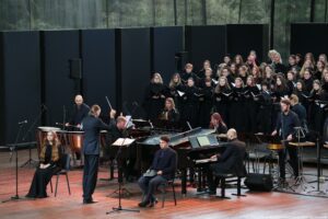 Ujęcie bokiem. Scena. Na środku dyrygent (bokiem). Po jego bokach ubrani na czarno siedzą chórzystka i chórzysta. Za nimi orkiestra, a za nią stoi ubrany na czarno chór – ujęcie na około 40 osób.