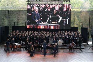 Scena. Na środku dyrygent (tyłem). Po jego bokach ubrani na czarno siedzą chórzystka (po lewej) i chórzysta. W tle orkiestra, a za nią stoi ubrany na czarno chór – ponad 100 osób. Nad nimi duży ekran prezentujący występ.