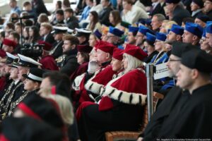 Ujęcie bokiem. Inauguracja roku akademickiego. Siedząca widownia, w tym osoby w mundurach i togach z granatowymi elementami oraz prorektorzy w czerwonych togach z gronostajami.