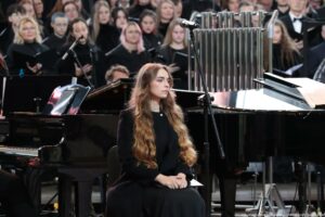 Scena. Po lewej siedząca bokiem chórzystka ubrana na czarno, za nią czarne fortepiany. W tle ubrany na czarno chór.