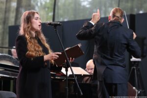 Scena. Po lewej śpiewająca chórzystka (bokiem), po lewej dyrygent z batutą (tyłem). W tle fortepian i pianista.