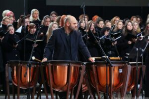 Scena. Na pierwszym planie cztery bębny i bębniarz. W tle ubrani na czarno chórzyści trzymający nuty.