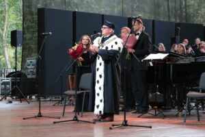 Na scenie przy mikrofonie stoi ubrany w czarną togę z gronostajami rektor Zachodniopomorskiego Uniwersytetu Technologicznego. Za nim troje muzyków z kwiatami (kobieta i dwóch mężczyzn). W tle fortepian i chórzyści.