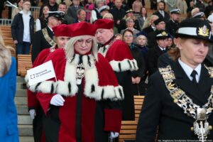 Po schodach Teatru Letniego schodzą ubranie w czerwone togi z gronostajami prorektorzy Uniwersytetu Szczecińskiego (dwie kobiety i mężczyzna). Prorektor ds. kształcenia trzyma małą białą flagą z logiem Spacerku na uniwerek. Po prawej stronie władze innych uczelni i gości, którzy zajmują miejsca przed uroczystością inauguracji.