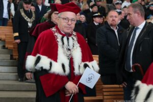 Po schodach Teatru Letniego schodzi ubrany w czerwoną togę z gronostajami prorektor Uniwersytetu Szczecińskiego trzymający małą białą flagą z logiem US. Po prawej stronie władze innych uczelni i oficjele, którzy zajmują miejsca przed uroczystością inauguracji.